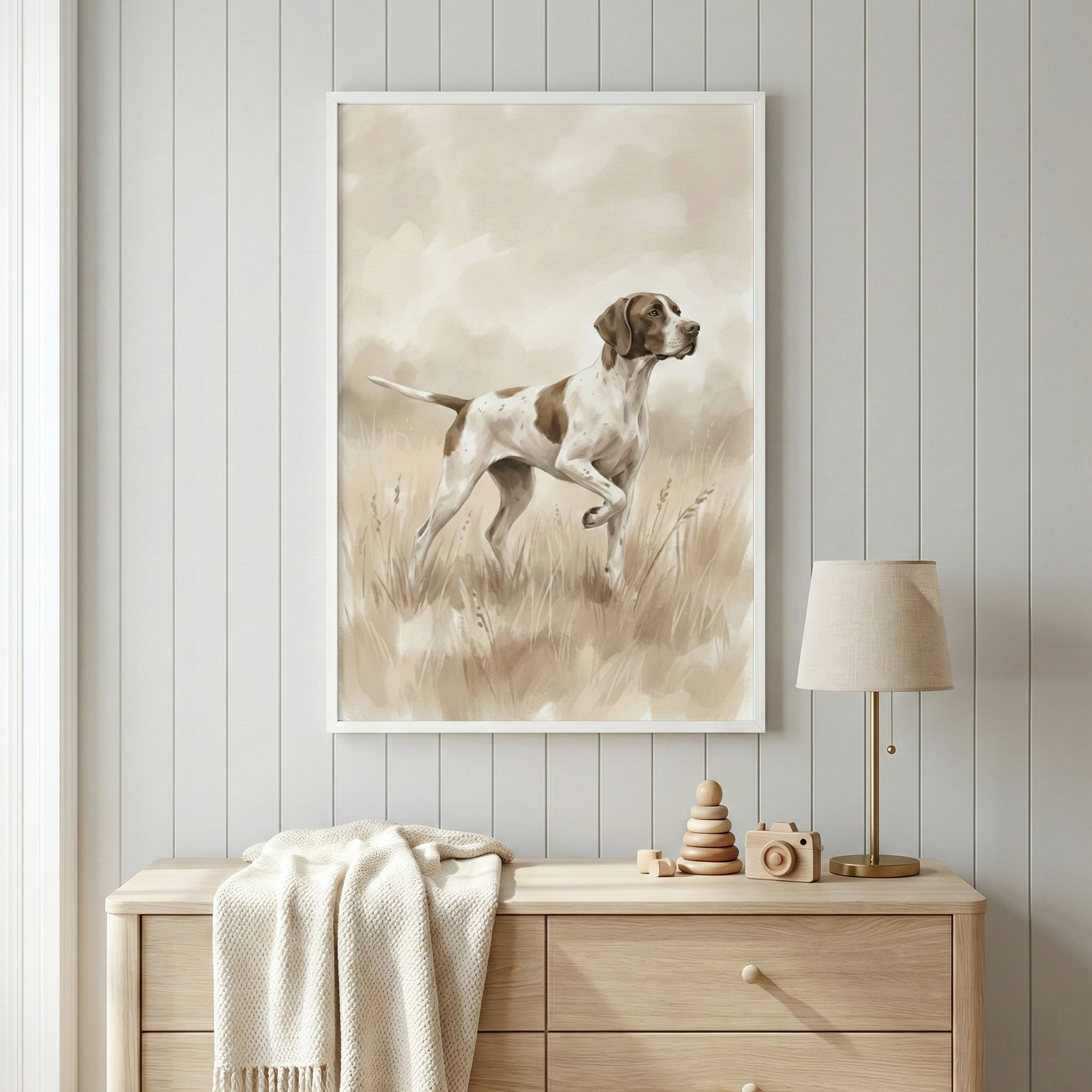 A framed artwork of a brown and white pointer dog in a field of tall grass hangs on a light grey vertical paneled wall. The illustration has a soft, muted color palette. Below the frame sits a light-toned wooden dresser. On top of the dresser, a cream-colored knit blanket is draped over one side. Next to it are a small wooden stacker toy, a wooden toy camera, and a gold table lamp with a beige shade. The scene has a clean, neutral, and minimalist aesthetic.
