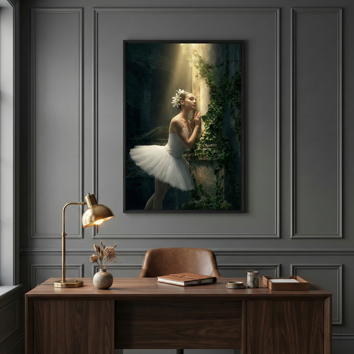 A sophisticated home office features dark grey paneled walls and a rich wood desk. Centered on the wall is a framed artwork of a ballerina in a white tutu, leaning against an ivy-draped stone pillar. She is bathed in a dramatic beam of light in an ethereal setting. Below, the desk holds a brass lamp, a leather notebook, and a small vase of dried flowers. A brown leather chair is tucked beneath, creating a moody and elegant workspace.