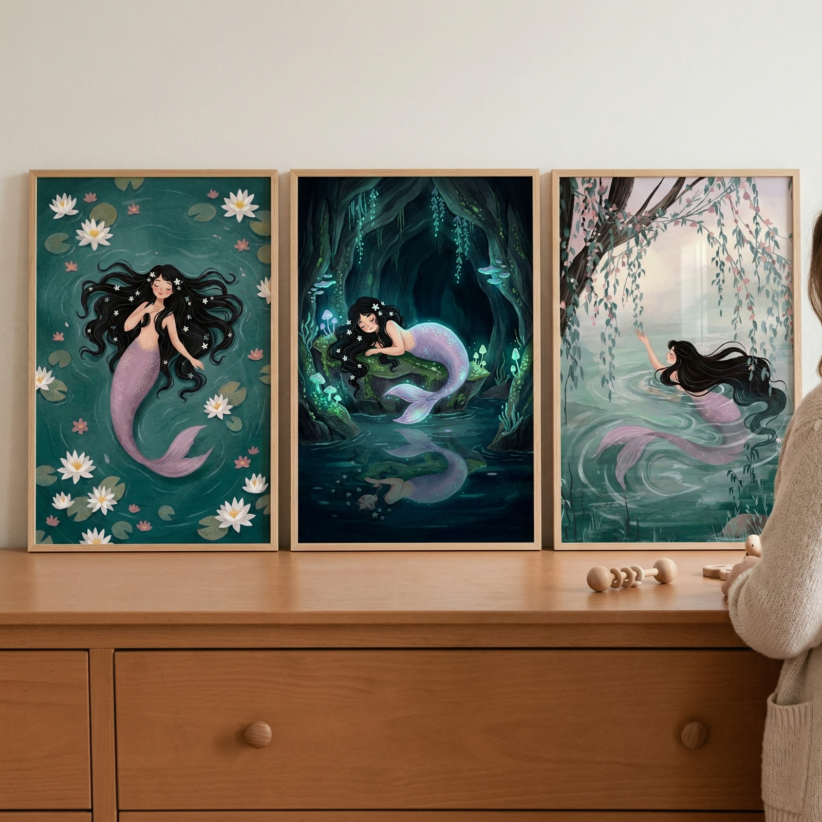 A set of three framed whimsical illustrations hang on a wall above a light wood dresser. Each artwork features a mermaid with long black hair and a lavender-pink tail in a different serene setting. The first shows her floating in deep green water among white water lilies. The second depicts her resting in a dark, bioluminescent cave. The third shows her swimming near a weeping willow tree. A wooden toy sits on the dresser, and a person's arm and sweater are partially visible on the right.