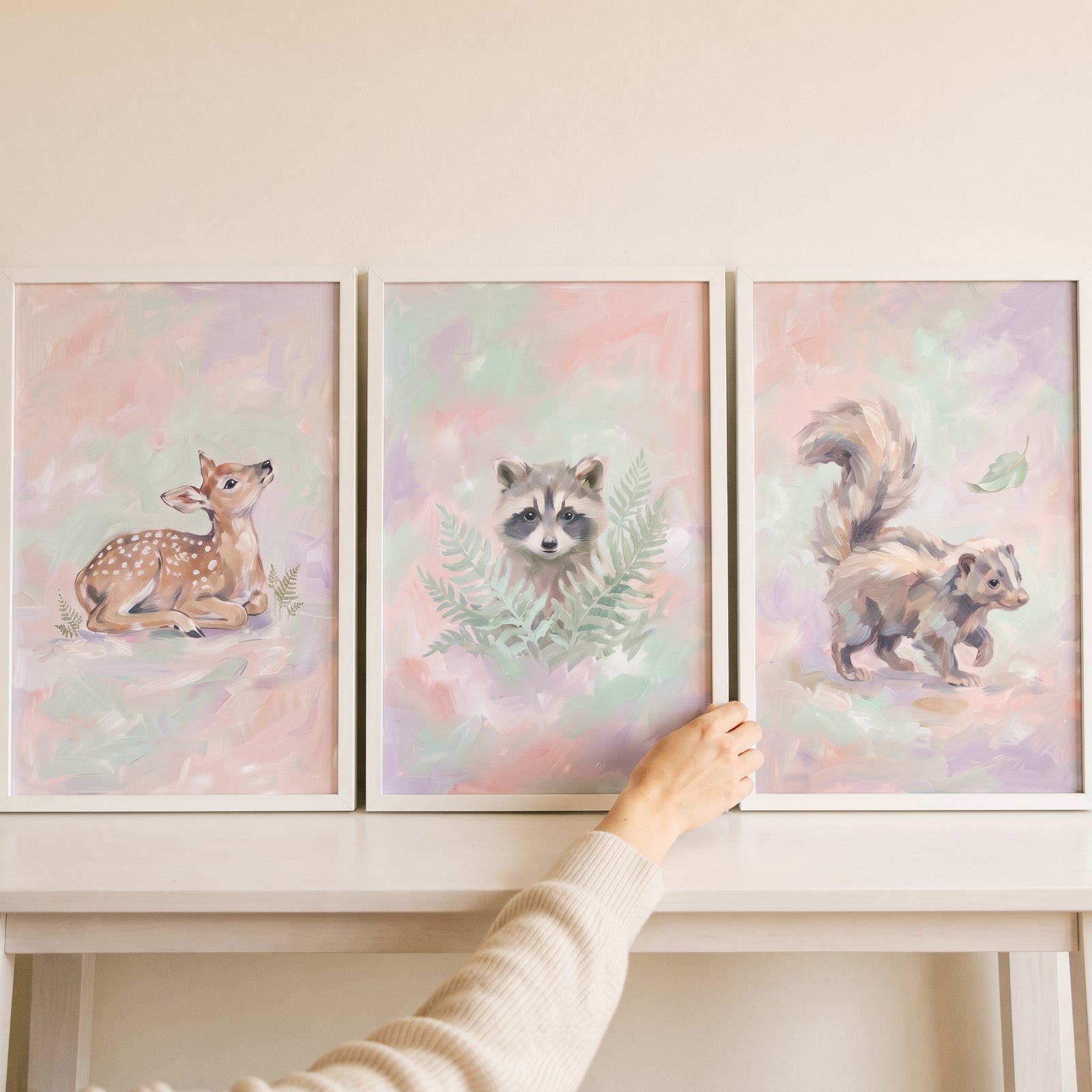 Hand adjusting an ethereal blush woodland animal wall art set of 3 showing a fawn, raccoon, and skunk in white frames.