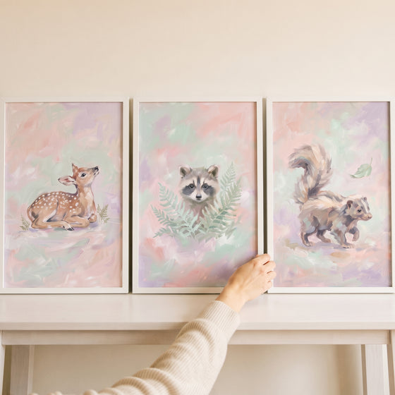 Hand adjusting an ethereal blush woodland animal wall art set of 3 showing a fawn, raccoon, and skunk in white frames.