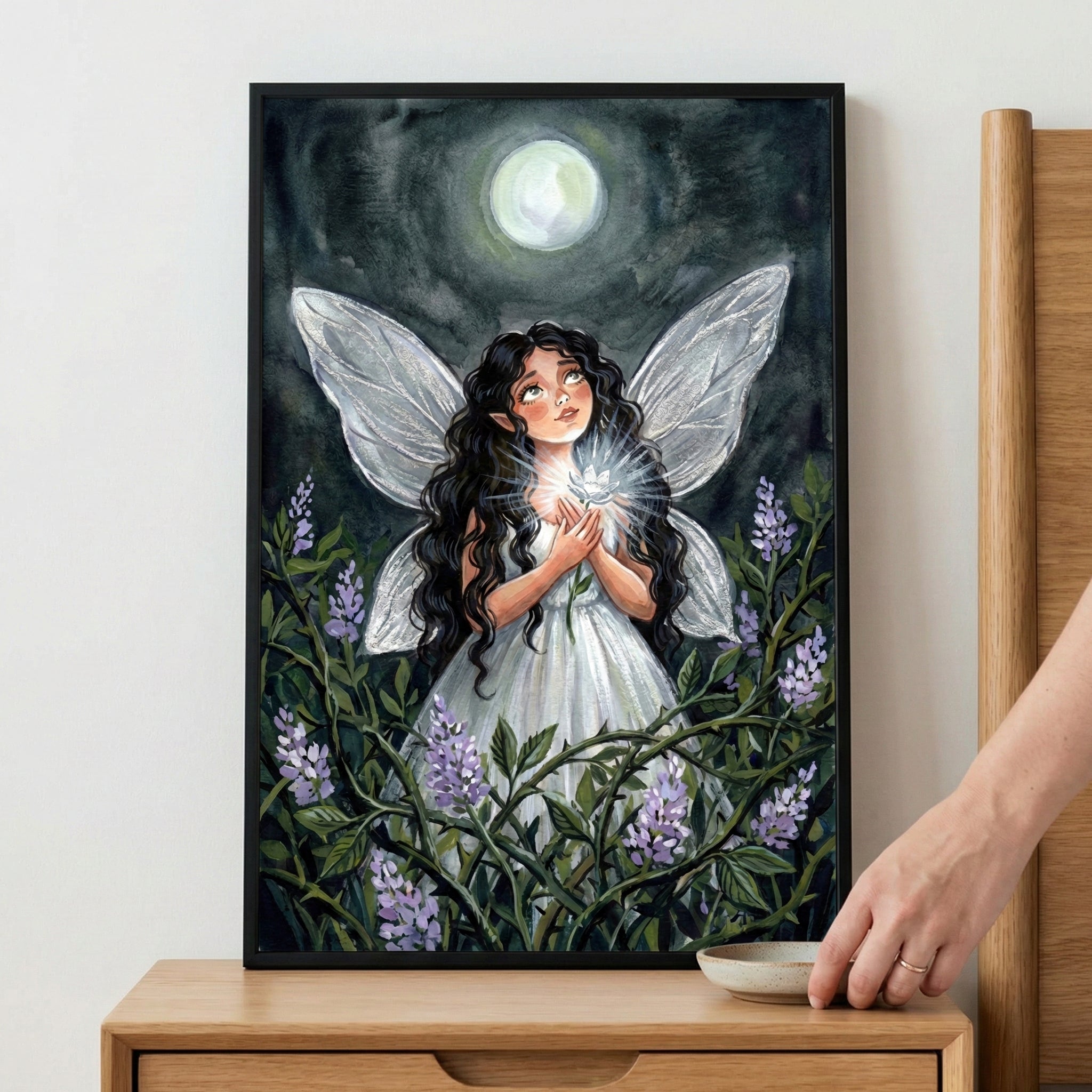 A framed whimsical illustration of a fairy-like figure with long dark hair, pointed ears, and large translucent wings. She is dressed in a simple white gown and stands amidst green leaves and purple blossoms, holding a small glowing light in her hands. She gazes upwards towards a bright, full moon in a dark, textured sky. The artwork is displayed in a black frame on a wooden surface.