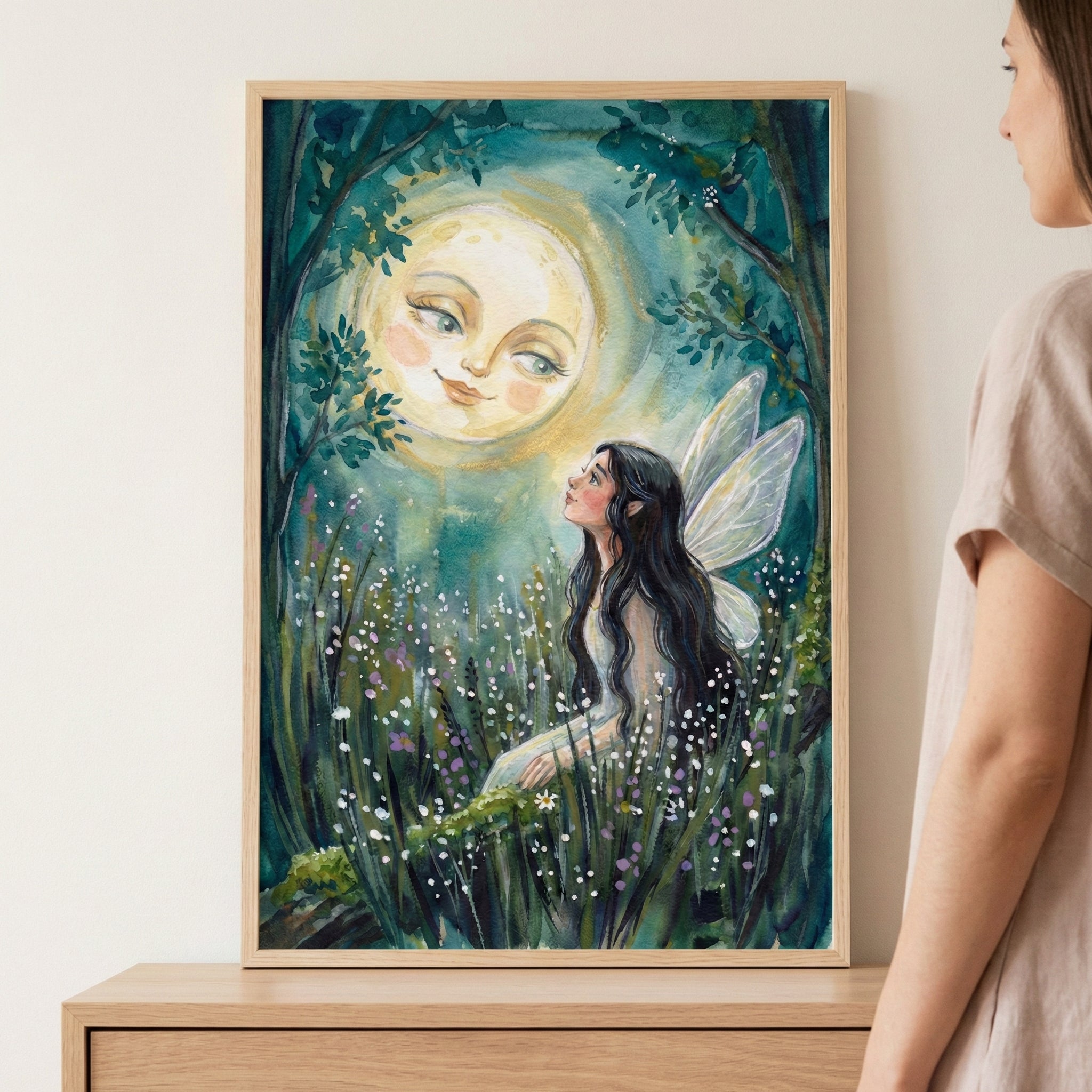 A framed vertical illustration depicting a whimsical night scene. A woman with long dark hair and delicate, translucent wings sits amidst tall grass and small white flowers, looking upward. Above her, a large, glowing full moon with a gentle, smiling human face illuminates the scene. The setting is a dark forest with leafy branches framing the top. The artwork, rendered in a soft illustrative style with deep greens, blues, and warm yellows, is displayed in a light wood frame resting on a wooden surface.