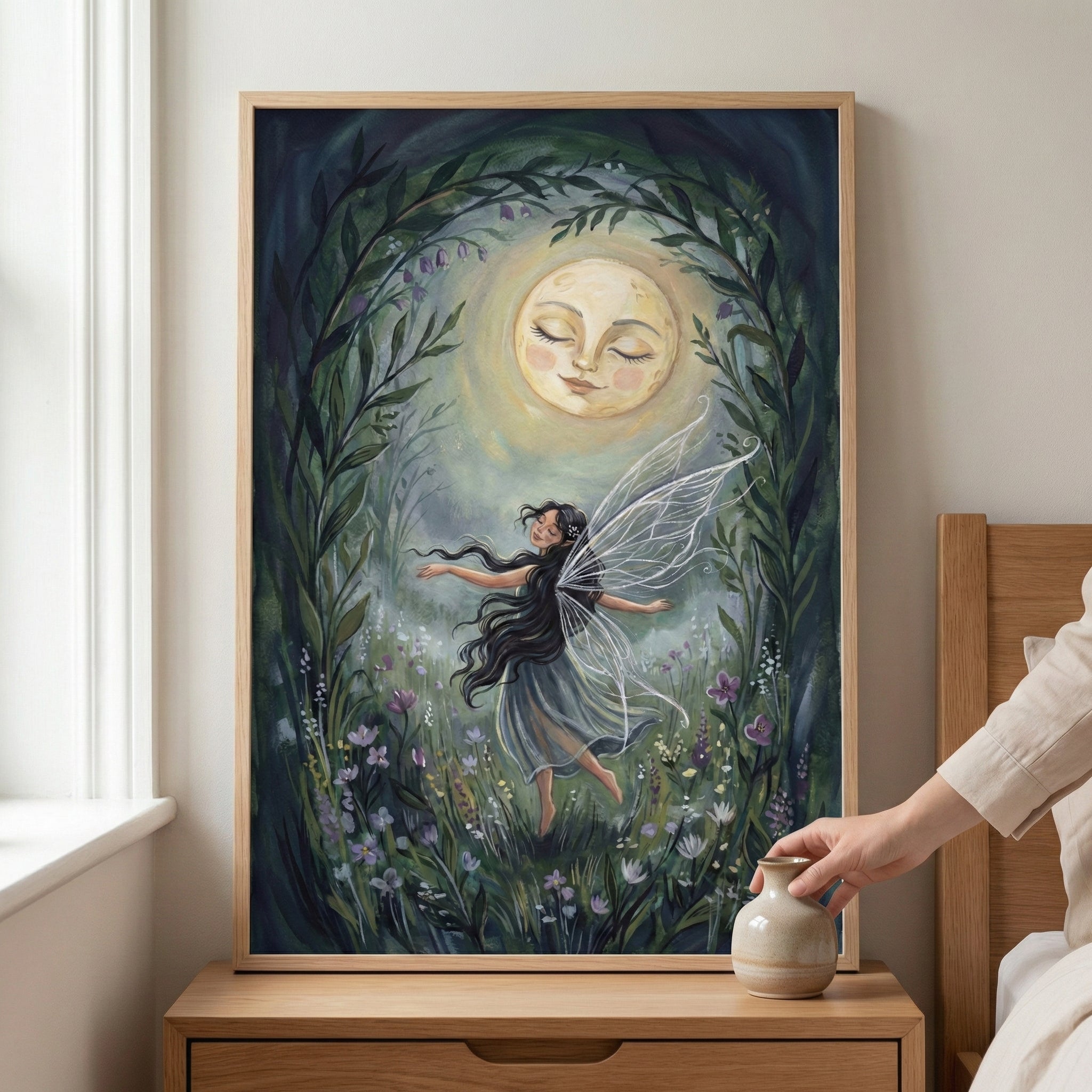 A framed illustration on a wooden nightstand featuring a fairy with long dark hair and shimmering translucent wings dancing amidst a field of purple and white wildflowers. Above her, a large, glowing full moon with a peaceful human face shines down, all framed by an archway of dark green leaves. To the right, a person's hand reaches for a small ceramic vase.