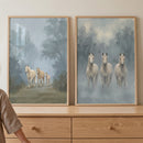 Two framed horse prints lean on a light wood dresser, with part of a person visible at the left edge for scale. One print shows three pale horses walking through a misty forest path, while the other shows three white horses standing side by side against a soft blue gray background.