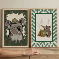 Two framed koala art prints are displayed on a wooden cabinet with a hand in front for scale. The left print shows a close up koala wearing a safari hat and surrounded by evergreen branches and red berries while the right print shows a smaller koala looking through a telescope beside a green tent marked "01" which highlights the playful woodland style of the set.
