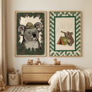 Two framed illustrations of koalas hang on a cream-colored wall above a light wood dresser in a child's nursery. The left illustration is a portrait of a koala wearing a tan safari hat, framed by evergreen branches and small red berries. The right illustration depicts a koala looking through a telescope next to a green tent labeled "01," set within a green and evergreen pattern border. On the dresser sit several wooden toys, including a train and a duck,.