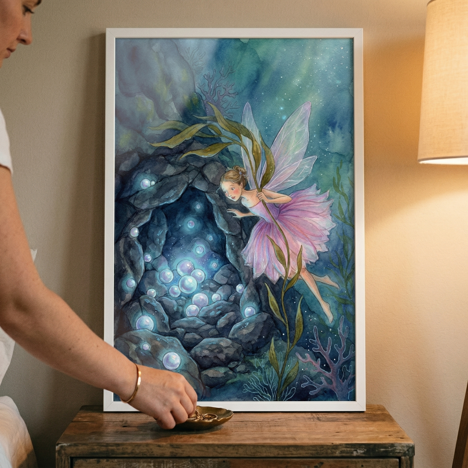 A framed watercolor-style illustration shows a magical underwater scene. A delicate figure with translucent, pale purple wings and a pink, ruffled skirt swims through deep blue and green water, holding a long strand of kelp. To her left, a dark, rocky cavern is filled with numerous glowing, iridescent orbs that cast a soft light. Detailed seaweed and coral pieces decorate the ocean floor. The artwork is held in a white frame and rests on a rustic wooden tabletop next to a warm lamp. 