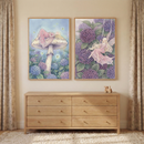 A cozy room features two large vertical framed artworks on a neutral wall. The left piece shows a fairy with long blonde hair and wings resting atop a giant mushroom, surrounded by purple and blue flowers. The right piece shows a fairy sitting on a swing nestled within dense purple blossoms. Below, a light-wood six-drawer dresser holds a small lamp with a soft glow and a wooden toy train. Long patterned curtains frame the scene, and a plush white rug covers the floor.