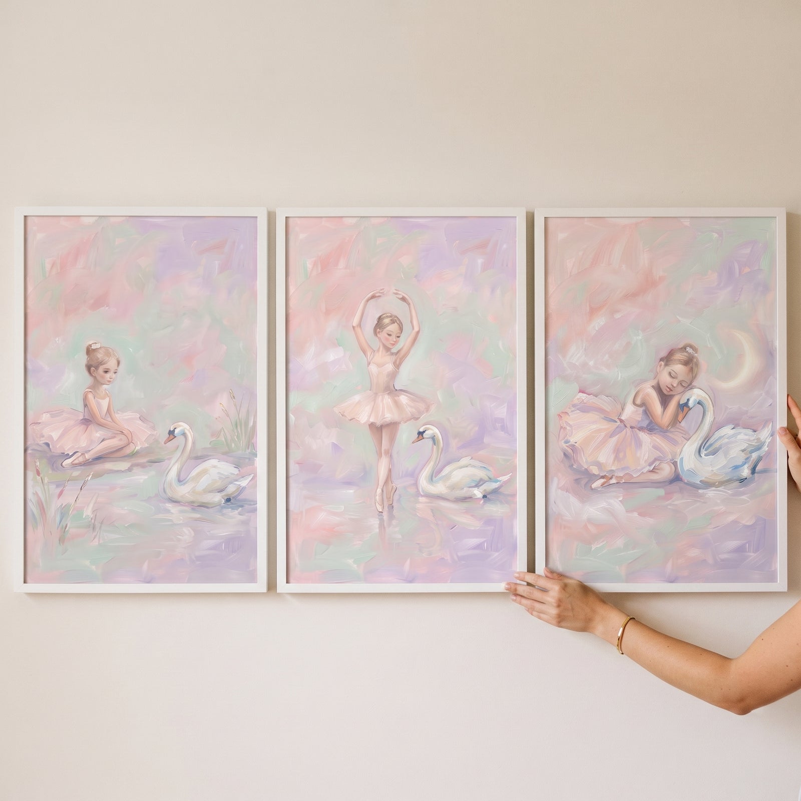 A hand adjusts a fairytale blush ballerina swan wall art set of 3 featuring a girl and swans in soft pink and purple hues.