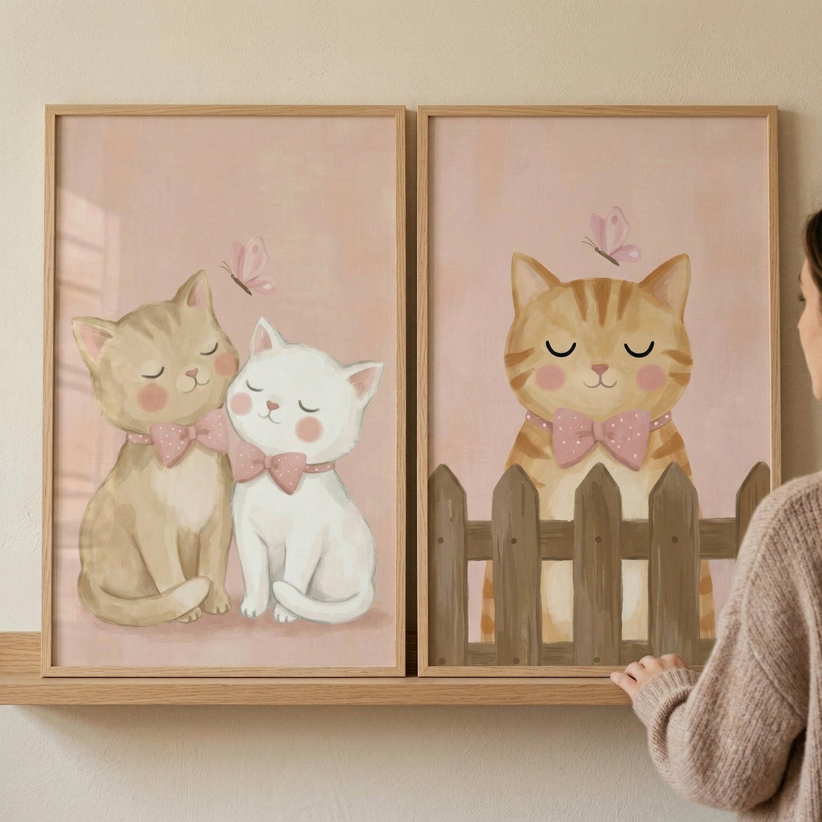Two vertical illustrations of whimsical kittens are displayed in matching light wood frames on a neutral-toned shelf. On the left, a light brown tabby cat and a white cat are depicted in a gentle embrace, both with closed eyes and soft pink cheeks. Each kitten wears a simple pink bow tie. A small, delicate pink butterfly is shown floating above them against a muted pink background. 
