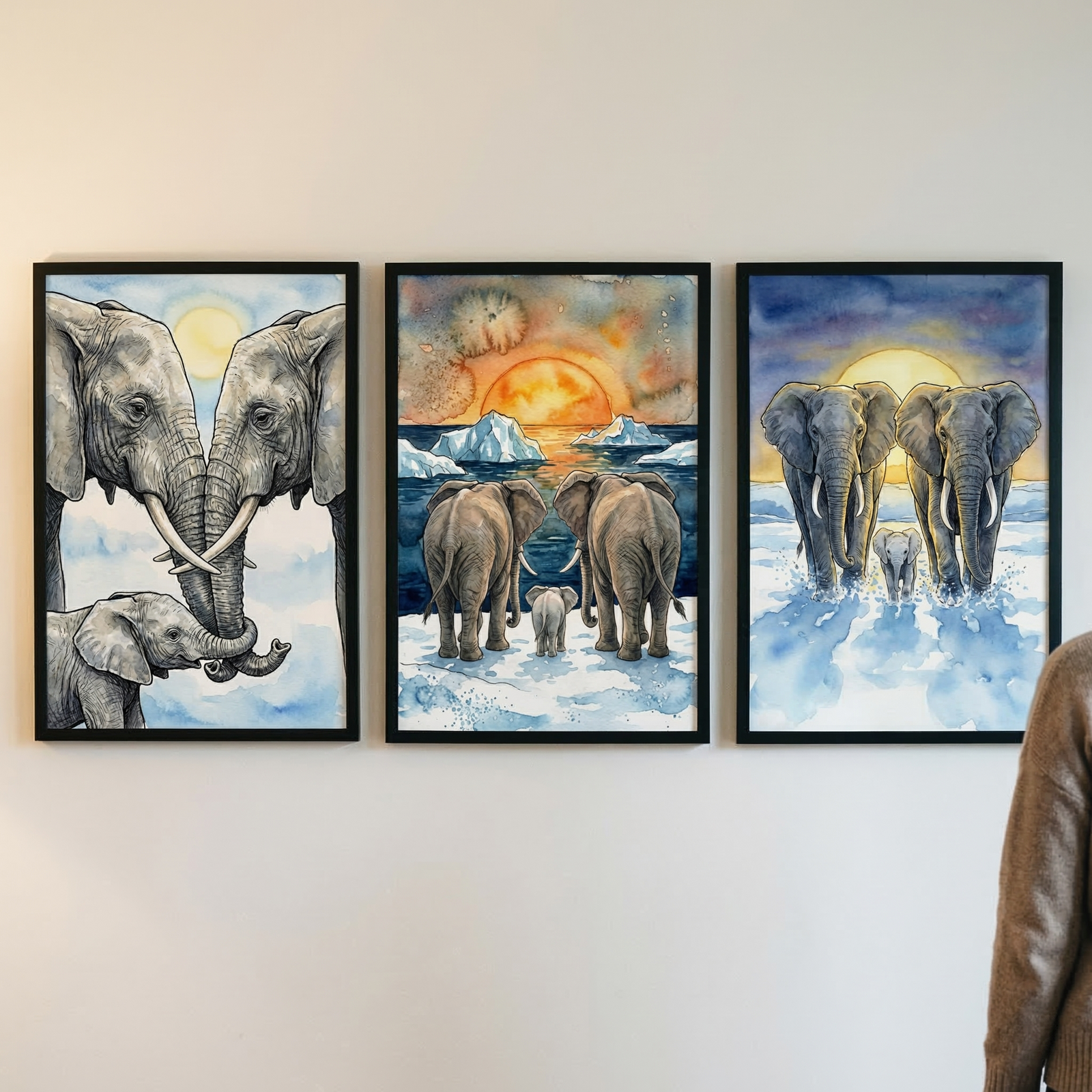 Three black-framed watercolor-style illustrations hang side-by-side on a plain white wall. Each piece features a family of elephants in a serene arctic environment. The left work shows two large elephants with trunks entwined over a calf. The center work shows the family from behind, looking at a large orange sun setting over icy water. The right work depicts the elephants walking forward through blue slushy water under a large yellow moon. A person's brown sweater is visible on the far right.
