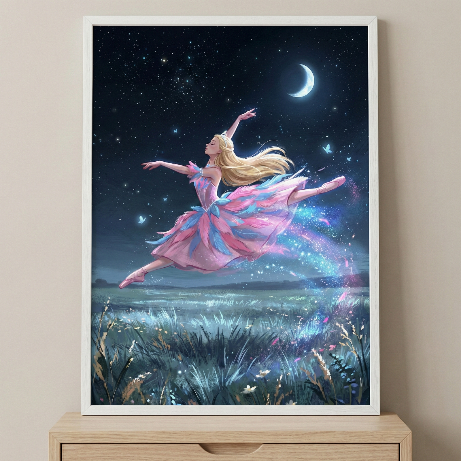 A framed illustration depicts a blonde ballerina leaping through a starry night sky. She wears a pink and blue tutu with a feathery texture and matching pink point shoes. A shimmering trail of sparkling light and small glowing butterflies follow her movement. Below, a field of tall grass is dimly lit under a bright crescent moon. The white-framed artwork is displayed on a light wooden cabinet against a neutral-colored wall.