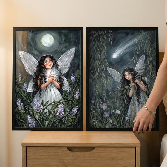 Two framed fantasy illustrations rest on a wooden nightstand. The image on the left features a dark-haired fairy with translucent white wings wearing a white dress. She holds a glowing light in her cupped hands, surrounded by purple flowers and dark greenery under a full moon. The image on the right shows a similar fairy peeking through willow leaves as a shooting star streaks across a dark sky. A person's hand is visible touching the side of the frame on the right.