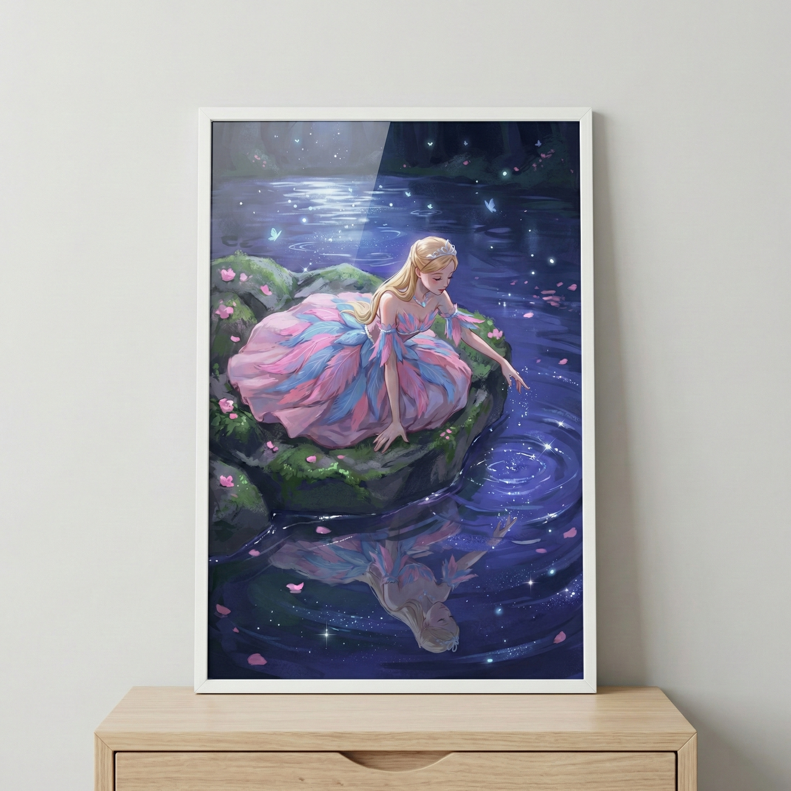 A framed fantasy illustration features a fair-haired young woman in a multi-layered pink and blue gown sitting on a mossy rock by a mystical pool at night. She reaches a hand toward the dark water, which ripples and glows with tiny shimmering lights. Her reflection is clearly visible beneath the surface. Glowing blue motes and butterflies fill the dark background. The artwork is held in a simple white frame, resting on a light wood surface against a neutral wall.
