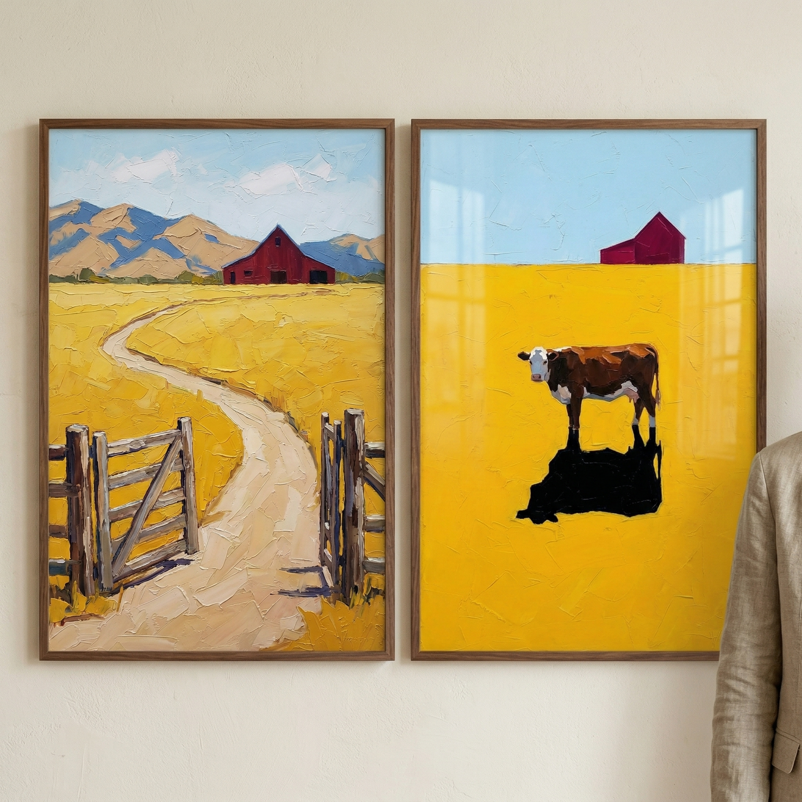 Two framed farm paintings hang side by side on a cream wall. The left painting shows an open gate and winding dirt road leading through yellow fields to a red barn and blue mountains. The right painting shows a brown and white cow standing alone in a bright yellow field with a red barn on the horizon. Part of a person in a beige jacket is visible at the right edge.
