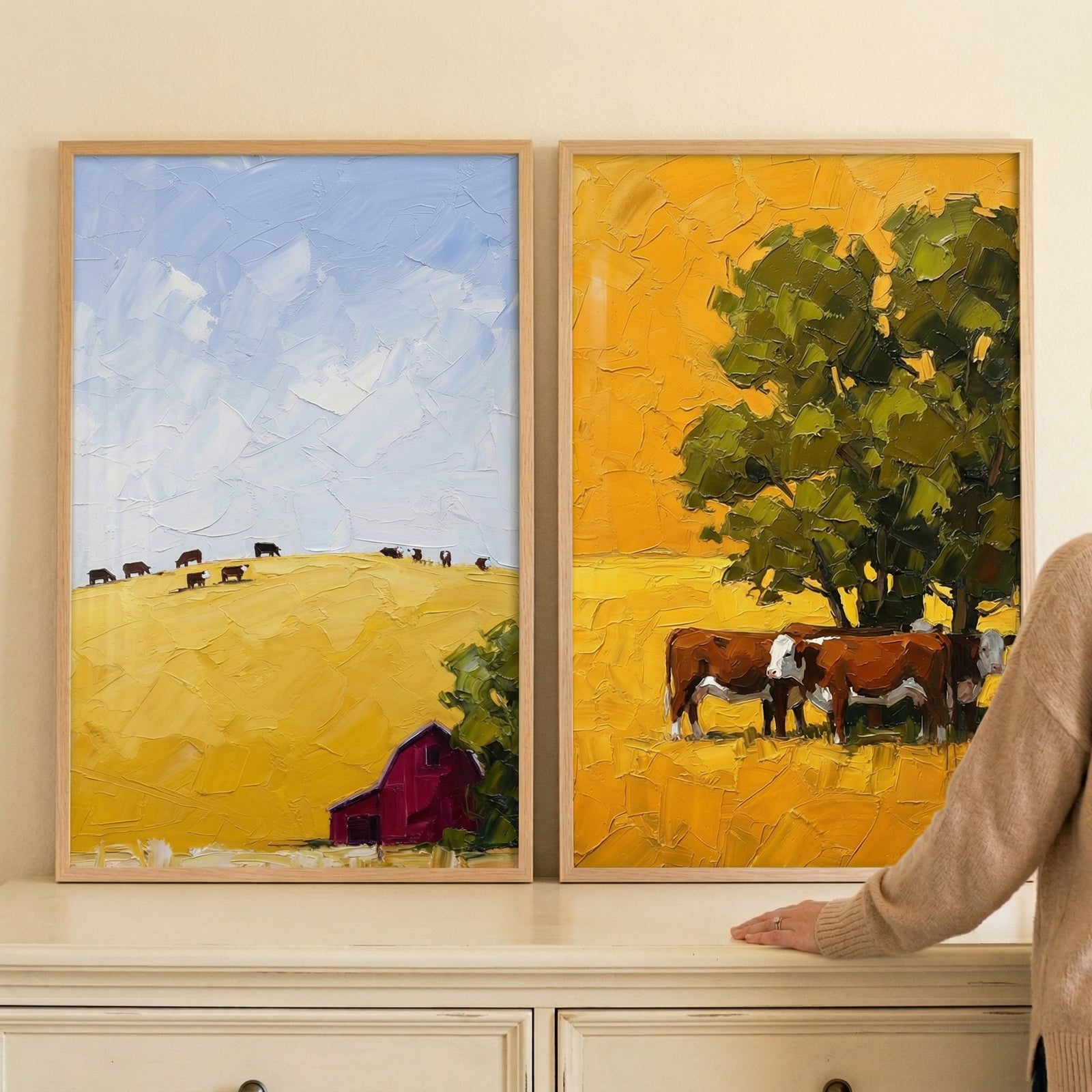 Two framed artistic creations are displayed side-by-side on a light wall above a white chest of drawers. Both pieces are rendered in a style characterized by thick, textured applications of color that create a rich, tactile surface.

The artwork on the left depicts a serene rural vista. A pale blue sky with textured white clouds hangs over a large, rolling yellow hill. Along the ridge of the hill, a row of small, dark animal silhouettes can be seen. 