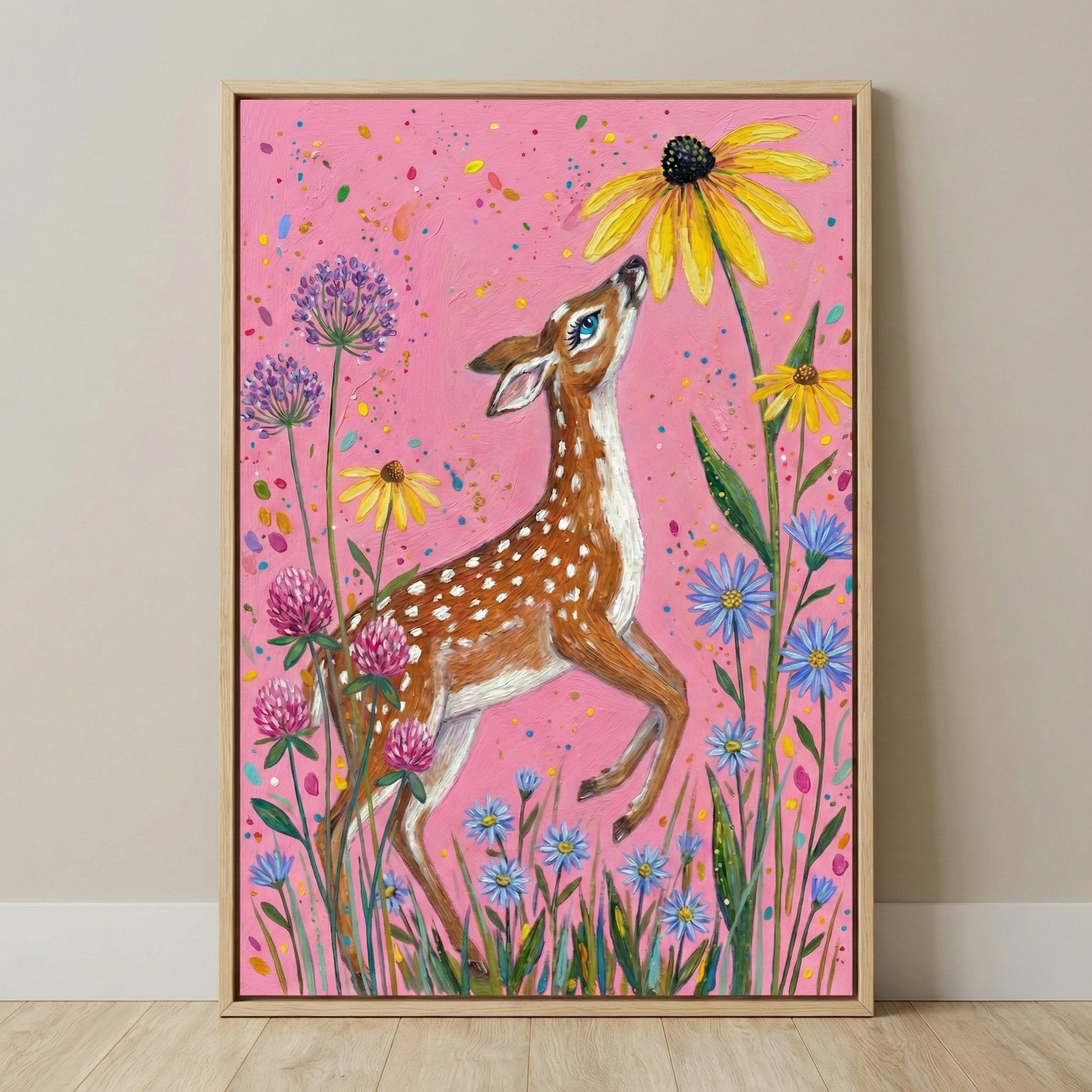 A whimsical vertical illustration features a young, spotted brown fawn reaching up to a large yellow flower. The fawn has expressive blue eyes and is surrounded by an array of colorful wildflowers, including blue, purple, and pink blooms. The entire scene is set against a bright pink background accented with multicolored splatters. This artwork is displayed in a thin, light-wood frame against a neutral wall and floor.