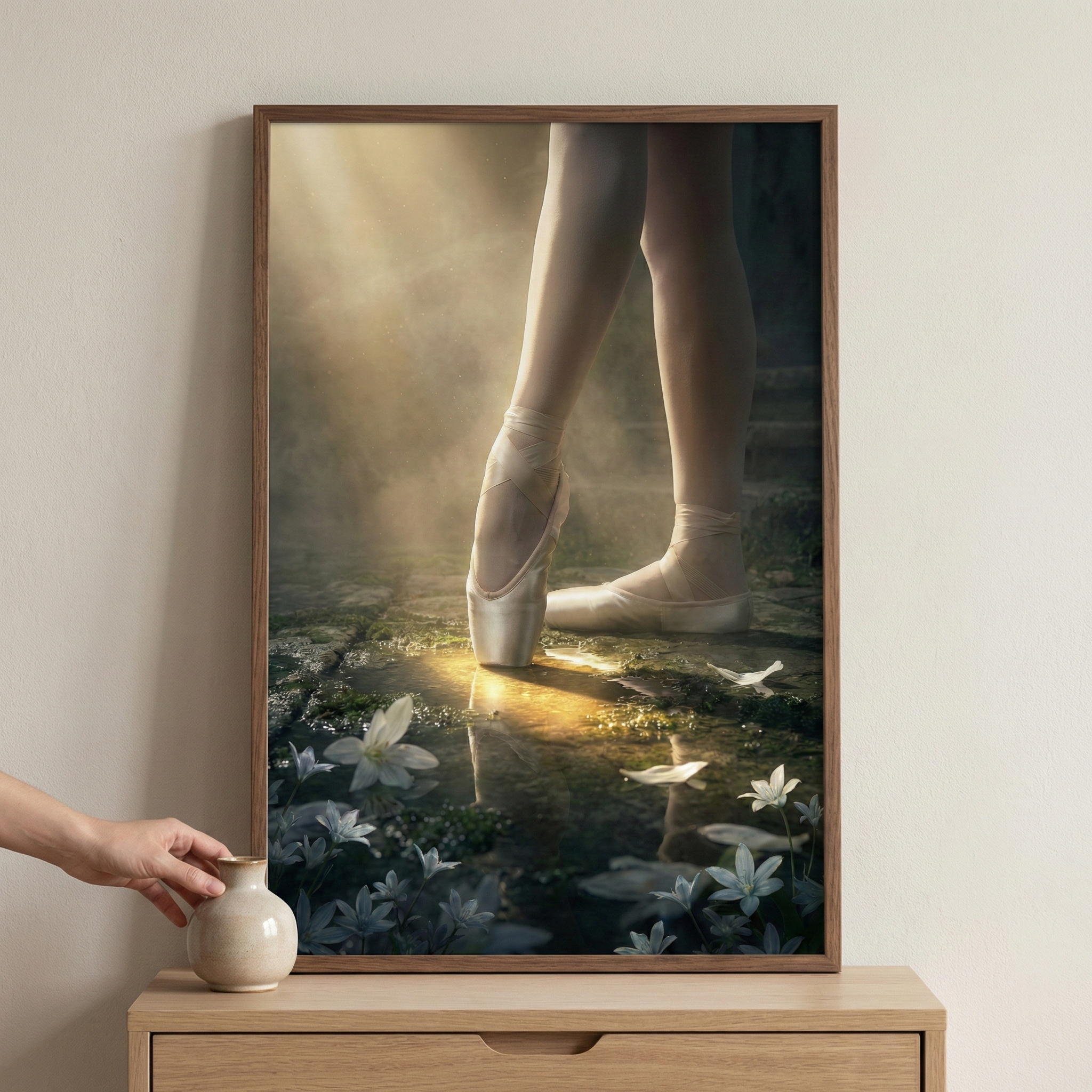 A framed artwork depicts the lower legs and feet of a ballet dancer. One foot is balanced on its tip in a satin slipper, while the other rests flat on a wet, stony ground. Delicate white flowers and petals are scattered around the dancer's feet. Soft beams of light break through a misty haze, creating a golden reflection on the floor. Below the frame, a hand reaches for a small ceramic vase resting on a wooden dresser. The entire image has a serene, dreamlike quality.
