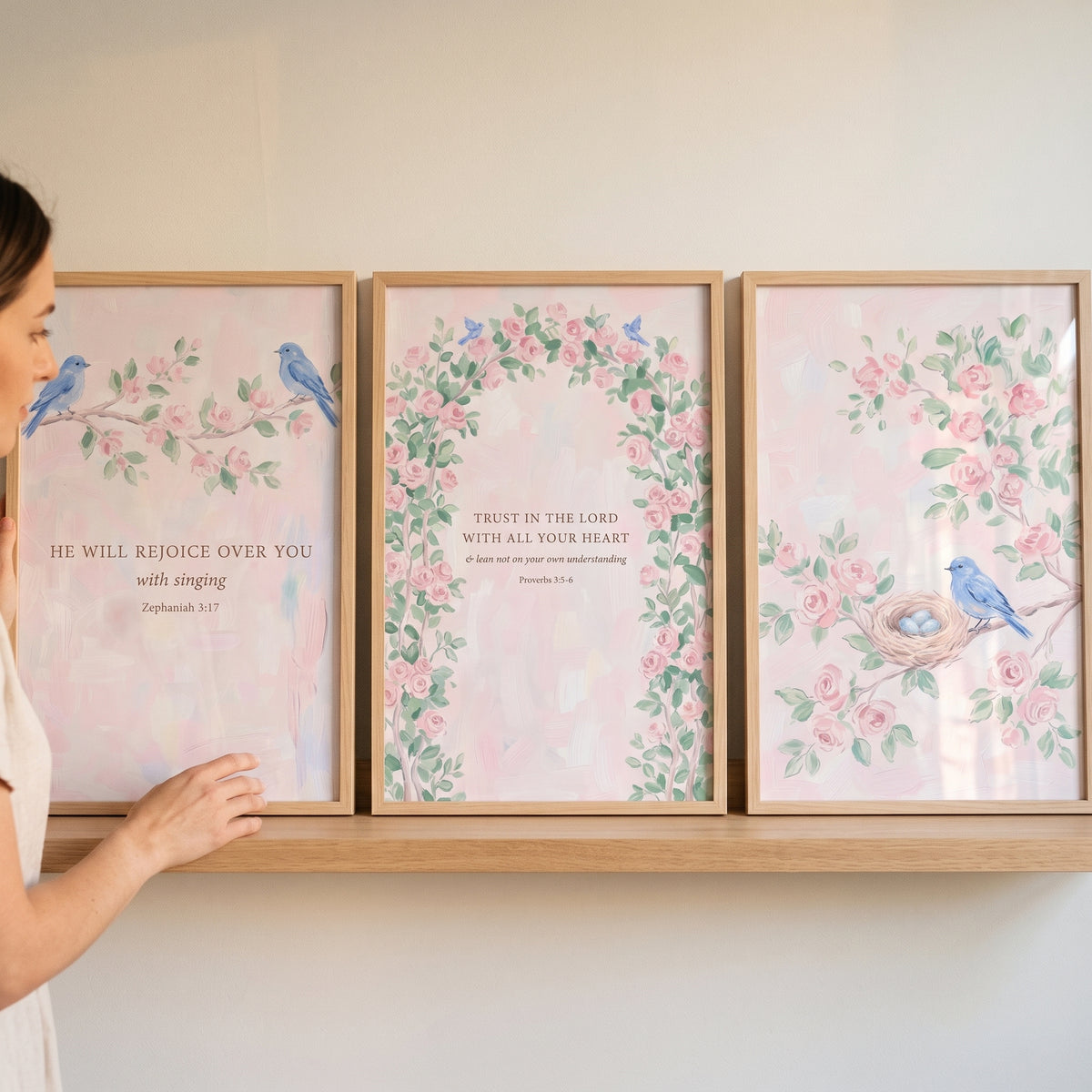 A woman arranges a feminine watercolor pink rose wall art set of 3 with blue birds and scripture on a shelf.