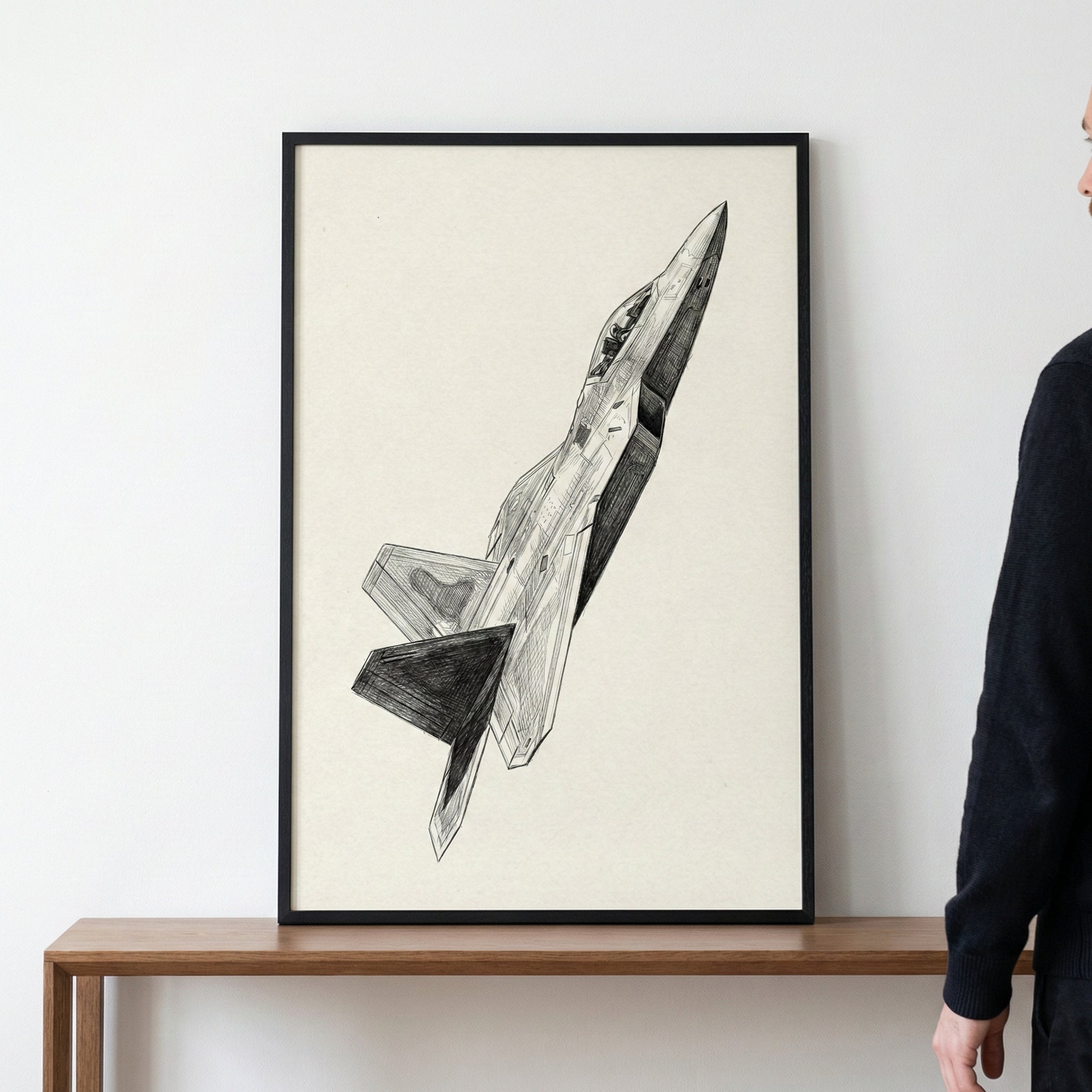 A framed black and white pencil sketch of a modern fighter jet soaring diagonally upwards. The detailed illustration features intricate shading and is presented on off-white paper within a thin black frame. The artwork sits on a wooden table against a plain white wall, with a person partially visible on the right.