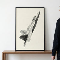 A framed black and white pencil sketch of a modern fighter jet soaring diagonally upwards. The detailed illustration features intricate shading and is presented on off-white paper within a thin black frame. The artwork sits on a wooden table against a plain white wall, with a person partially visible on the right.