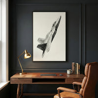 A sophisticated study area features a wooden desk set against a dark gray paneled wall. On the desk sits a gold-toned task lamp, a leather desk mat with a keyboard and mouse, a white mug, and a small stack of books. A large, black-framed vertical artwork hangs above the desk, displaying a detailed sketch of a modern fighter jet in flight. A brown leather office chair is tucked into the desk. To the left, the edge of a white-framed window is visible.