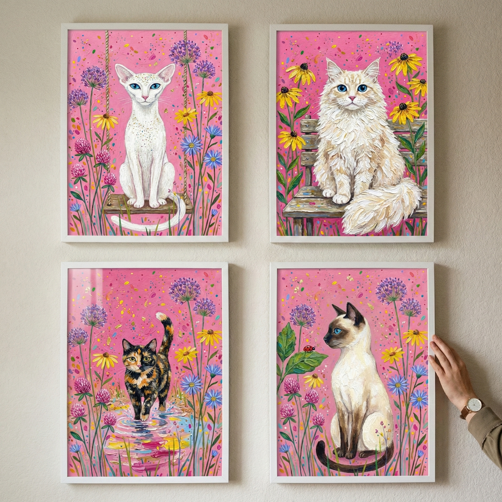 Four framed vertical artworks are arranged in a square grid on a plain wall. Each piece features a vibrant pink background decorated with colorful flowers. The top-left depicts a slender white cat sitting on a wooden swing. To its right, a fluffy cream cat rests on a weathered bench. Below, a tortoiseshell cat steps into a colorful puddle. The bottom-right features a sleek color-point cat sitting among purple blossoms. A person's hand reaches in to adjust the position of the bottom-right frame.
