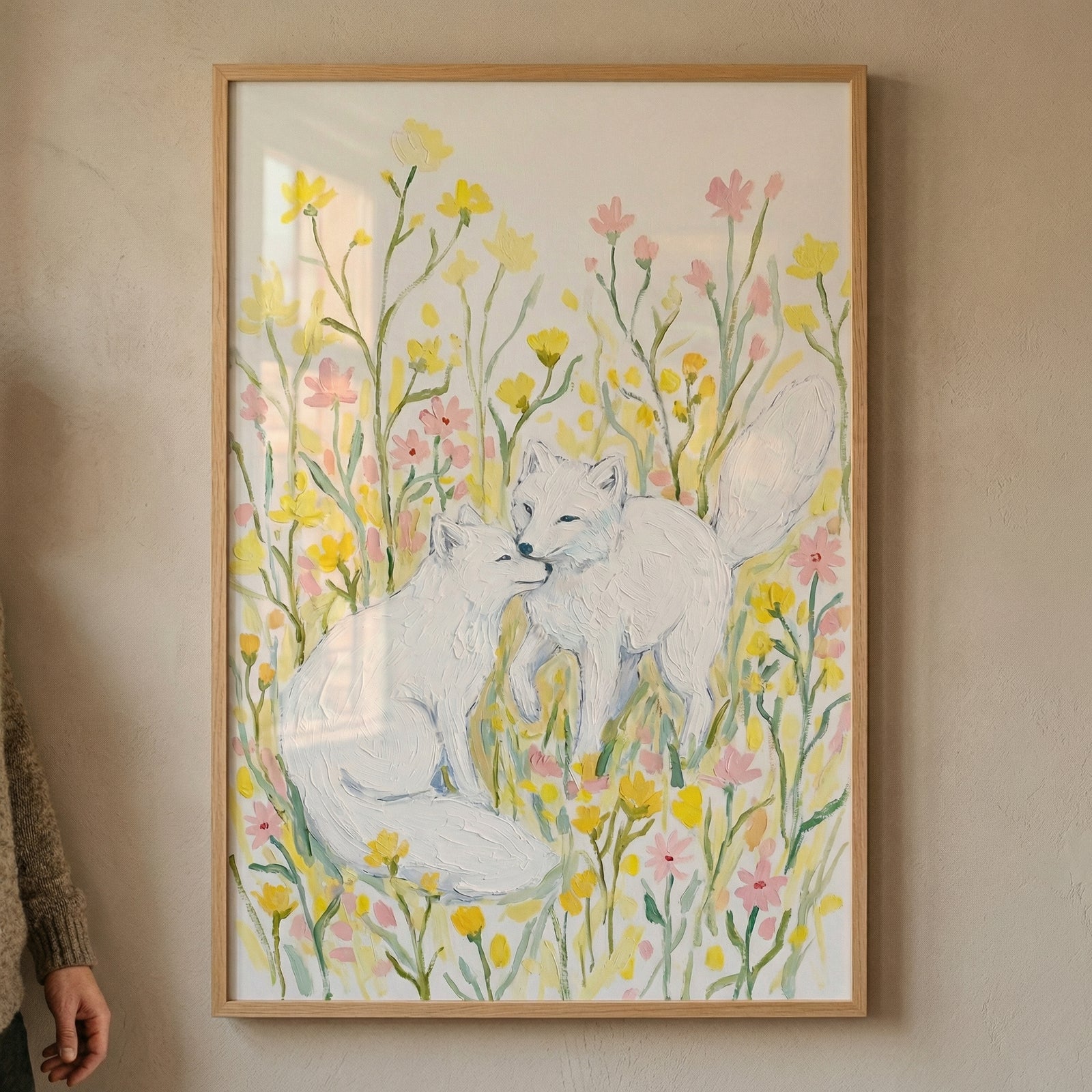 A framed illustration depicts two white foxes interacting within a field of yellow and pink wildflowers. One fox is seated while the other stands next to it, their noses touching in a gentle nuzzle. The artwork has a soft, textured appearance, with many green flower stalks and colorful blossoms against an off-white background. The piece is set in a light wooden frame and hangs on a neutral-colored wall. A person's hand is visible on the left edge of the scene.