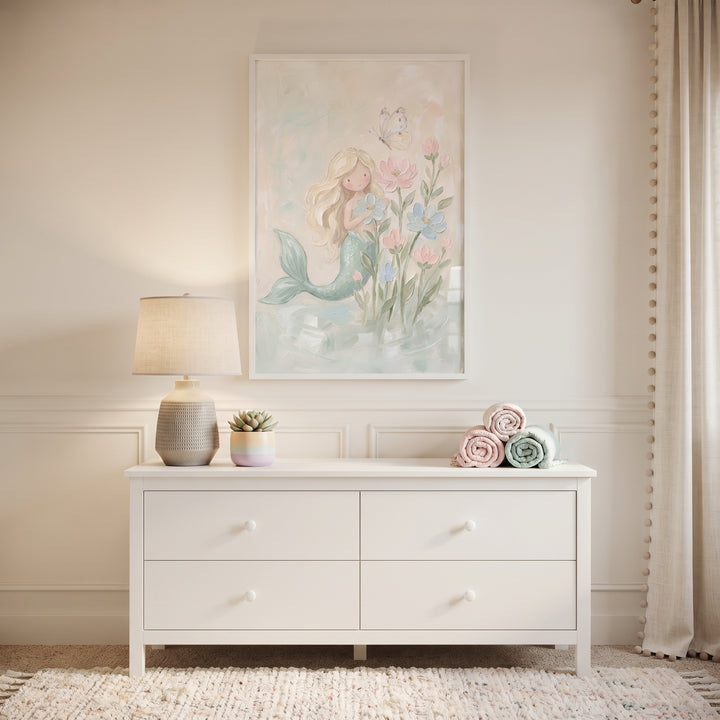 White dresser under floral pastel blonde mermaid wall art with a lamp, small plant, and rolled towels in a nursery.