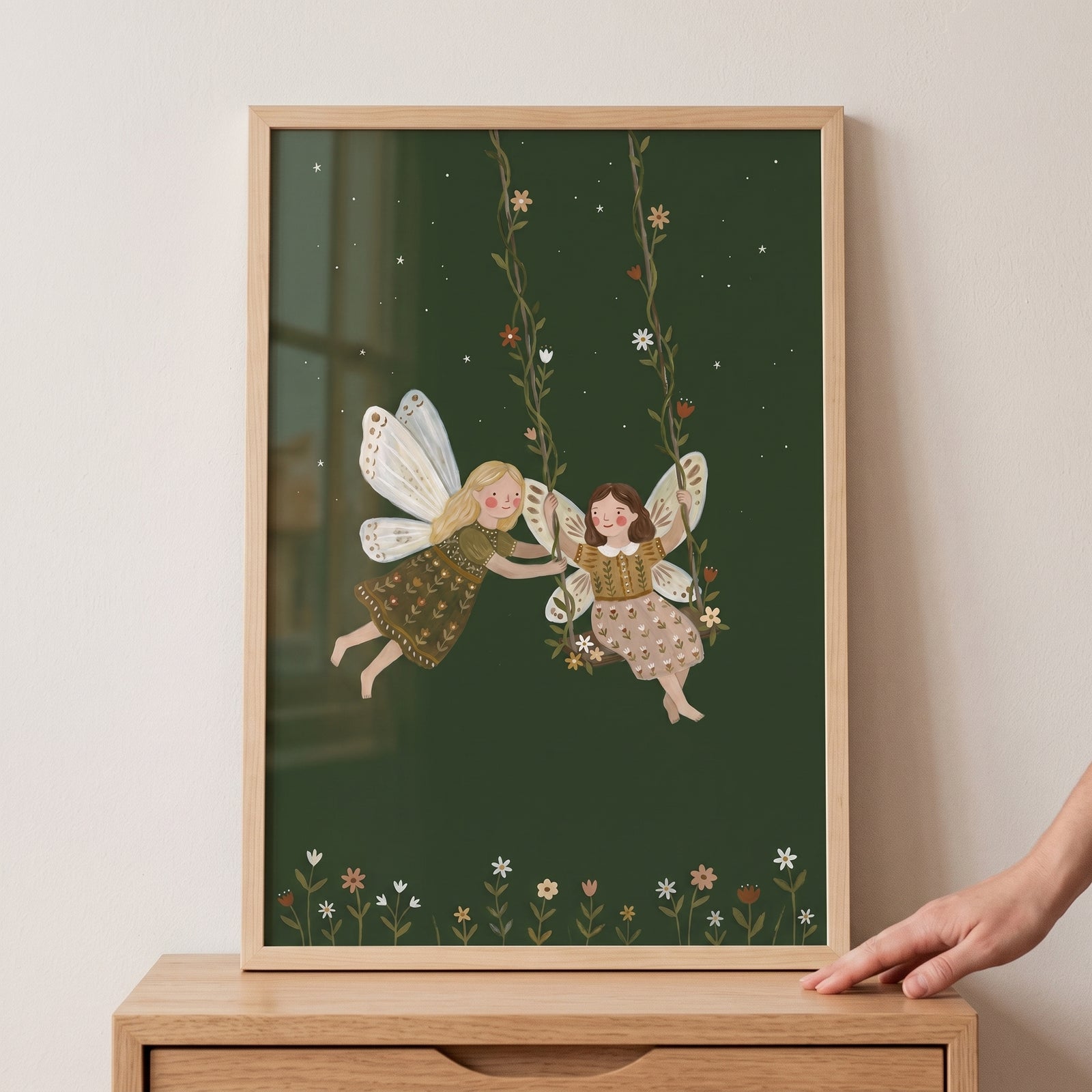 Floral vine swinging fairies wall art in a wood frame depicts two fairies on a vine swing against a dark green sky.