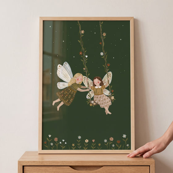 Floral vine swinging fairies wall art in a wood frame depicts two fairies on a vine swing against a dark green sky.