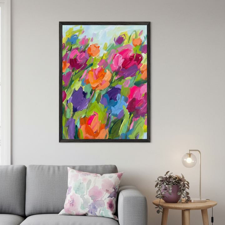 Modern living room with a gray sofa. A large black-framed abstract painting of vibrant, colorful flowers with bold brushstrokes hangs above the sofa. Colors include pink, orange, purple, blue, and green, with a light blue sky.