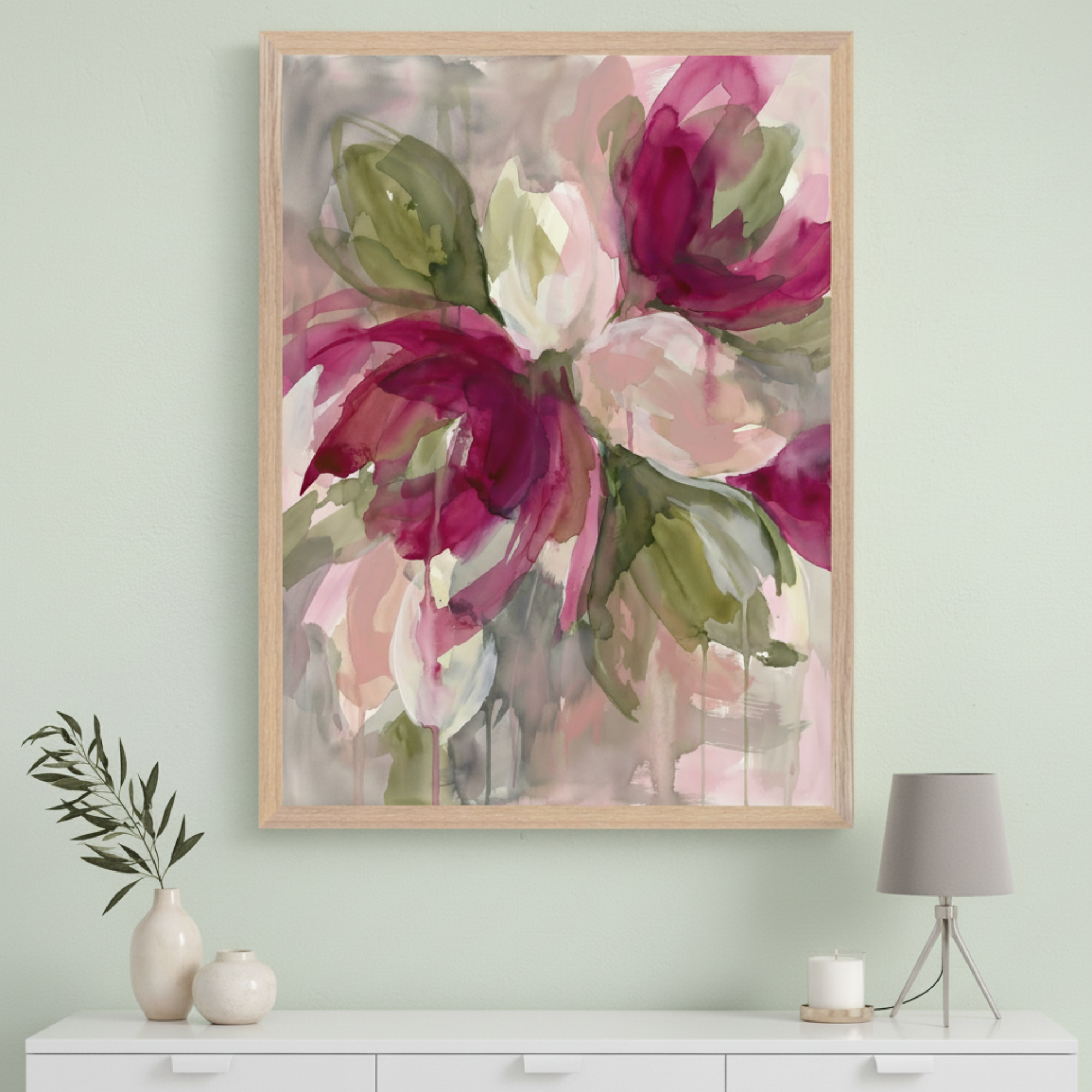 Modern room with a white dresser. A large light wood-framed abstract watercolor painting of magenta, pink, and olive floral shapes hangs centered above the dresser. Vases, a plant, a lamp, and a candle are on the dresser.