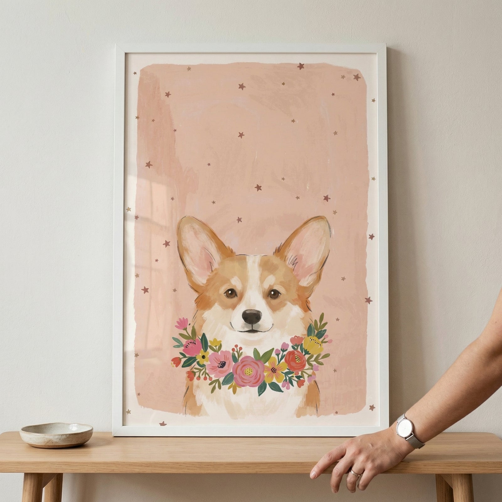 Framed illustration of a corgi with large upright ears wearing a colorful floral wreath around its neck against a soft peach pink background with scattered stars. A hand rests on the shelf beside the frame, adding scale and a warm styled nursery decor feel.