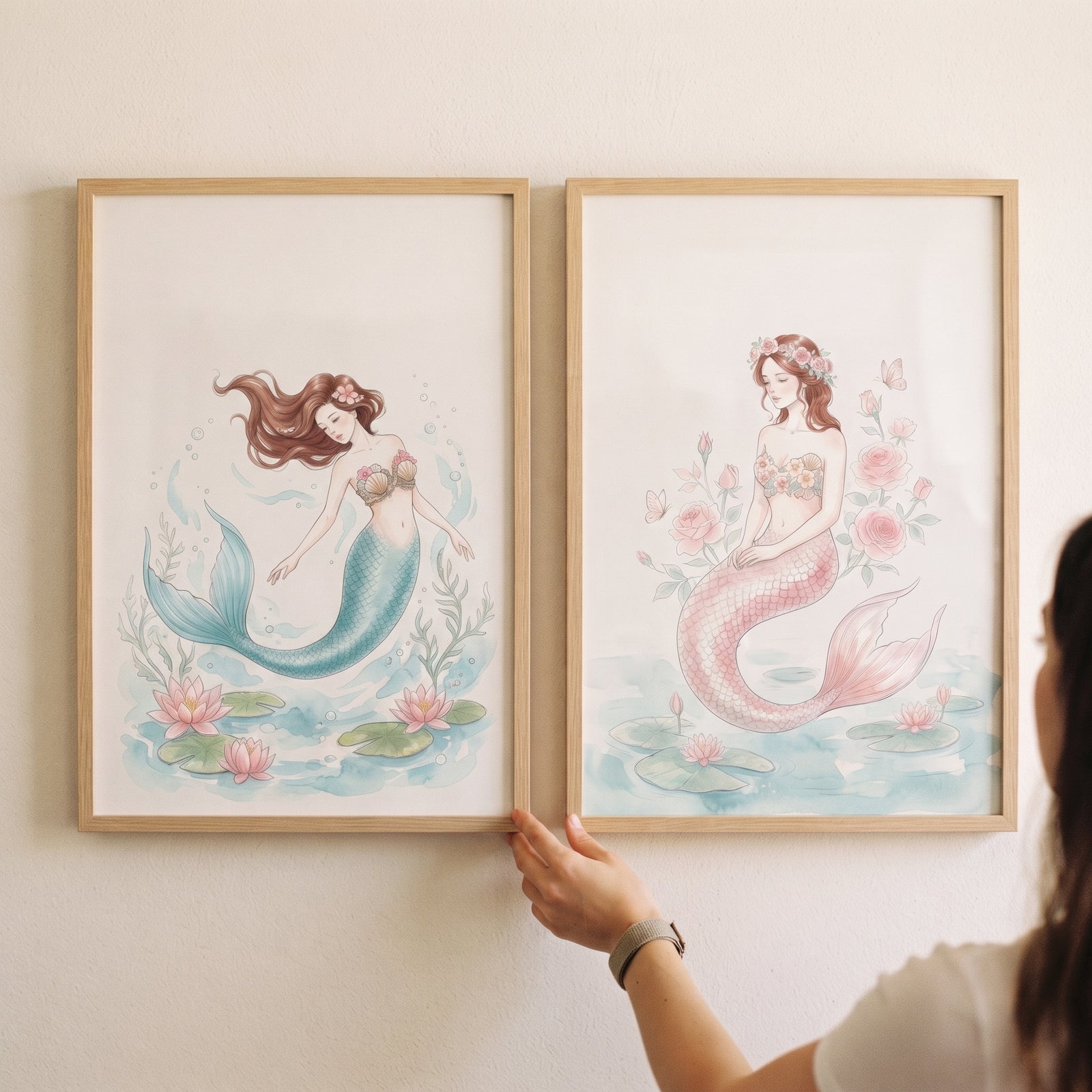 Hand touching a flowing auburn hair mermaid wall art set of 2 featuring watercolor mermaids with blue and pink tails.