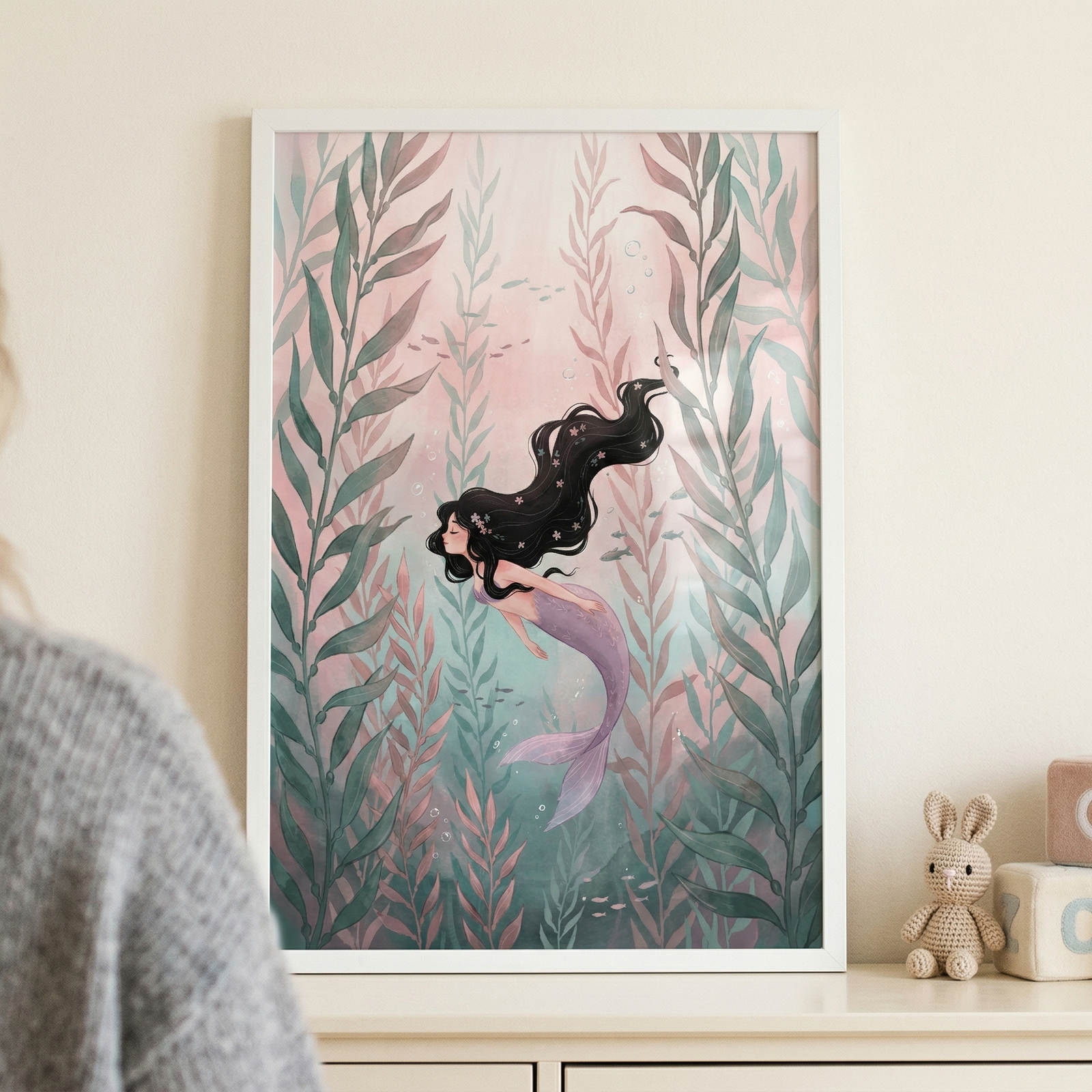 A vertical white frame holds an illustration of a mermaid swimming through an underwater scene. The mermaid has long, flowing dark hair and a purple tail, appearing to glide horizontally. She is surrounded by tall, leafy seaweed in soft shades of teal and muted pink. The background is a gentle, hazy pink with small bubbles and tiny fish scattered throughout. 