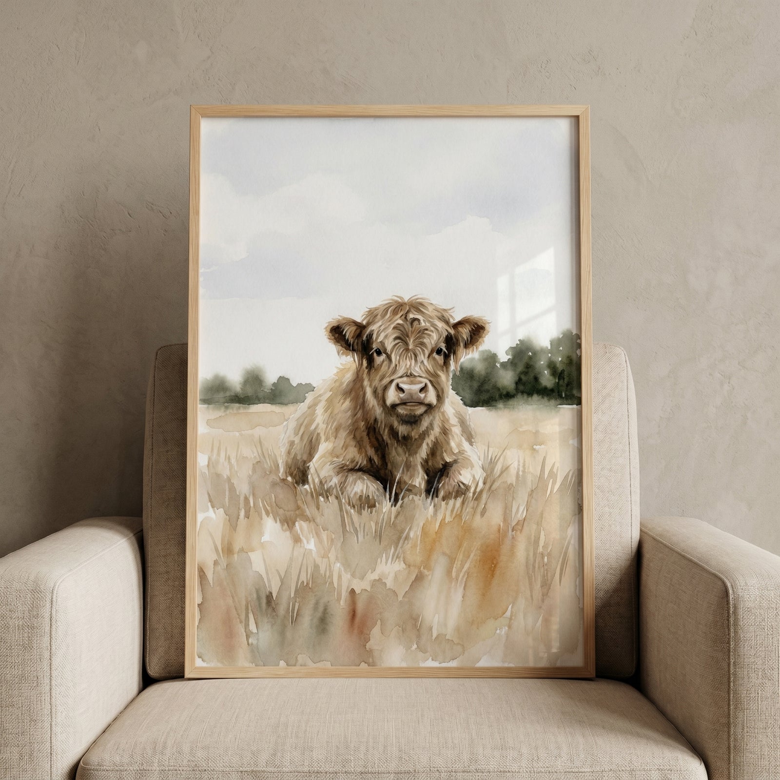 Framed fluffy baby highland cow wall art sits on a beige armchair. Soft watercolor style depicts the calf in a field.