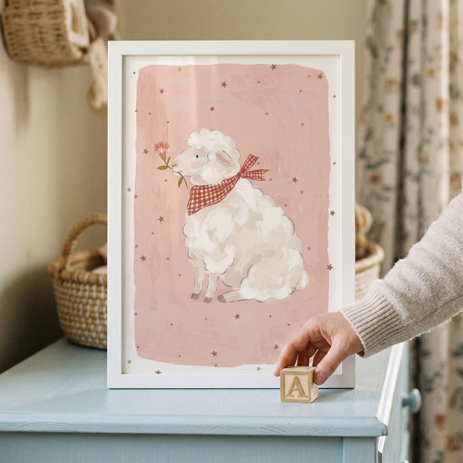 A framed vertical illustration of a fluffy white lamb is displayed on a light blue wooden dresser. The lamb, drawn in a whimsical style, wears a red and white checkered bow tie and holds a delicate pink flower in its mouth. The background of the artwork is a soft, muted pink decorated with small, dark pink stars. In the foreground, a person's hand places a wooden toy block featuring the letter "A" in front of the white frame. 