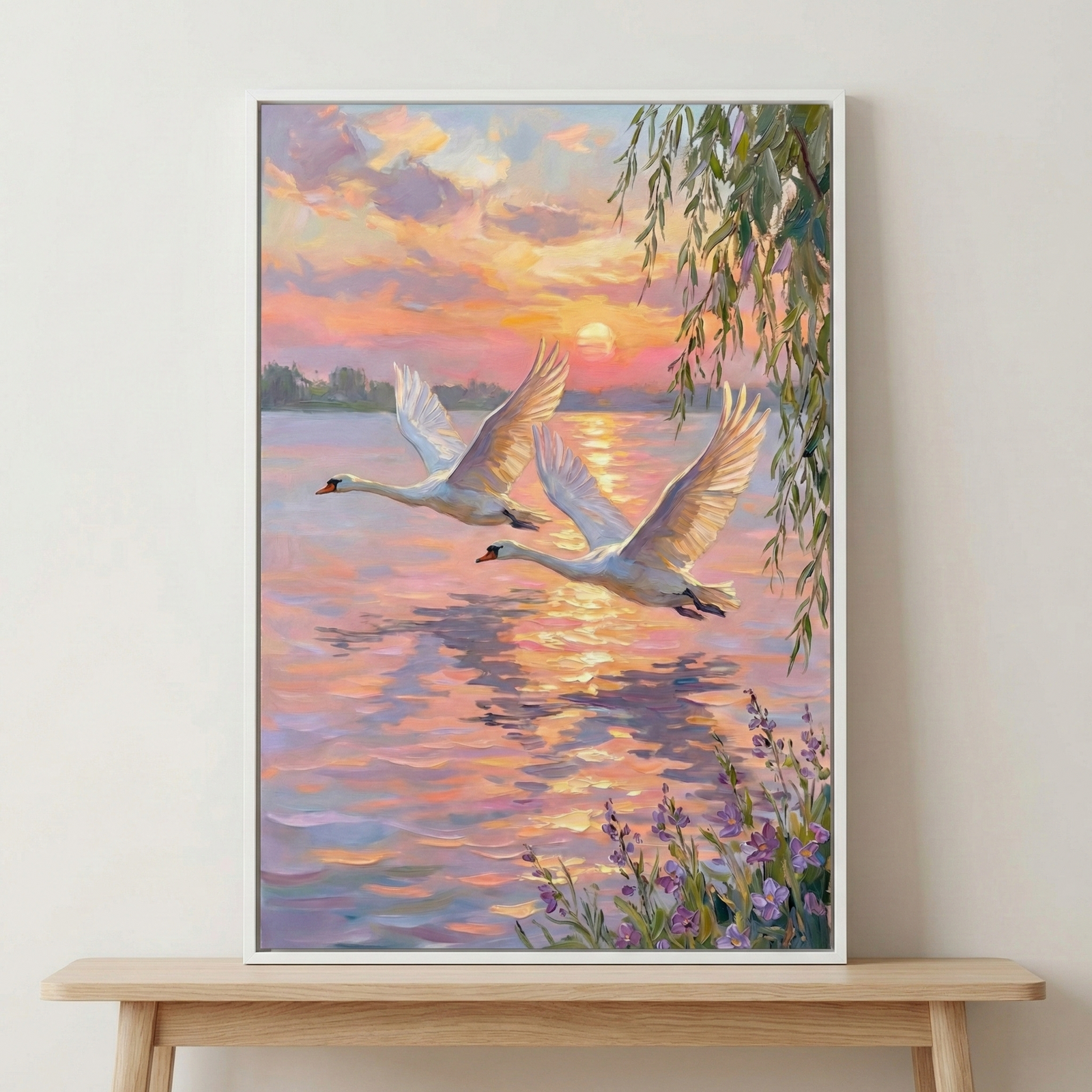 An impressionistic artwork features two white swans gliding above a peaceful lake during a radiant sunset. The sky is a canvas of warm oranges, soft pinks, and gentle purples, with a low sun casting a shimmering path across the water. Delicate willow leaves frame one side, while a cluster of purple wildflowers adds detail to the foreground. This vertical composition is presented in a clean white frame, perched on a light wood bench against a neutral wall.