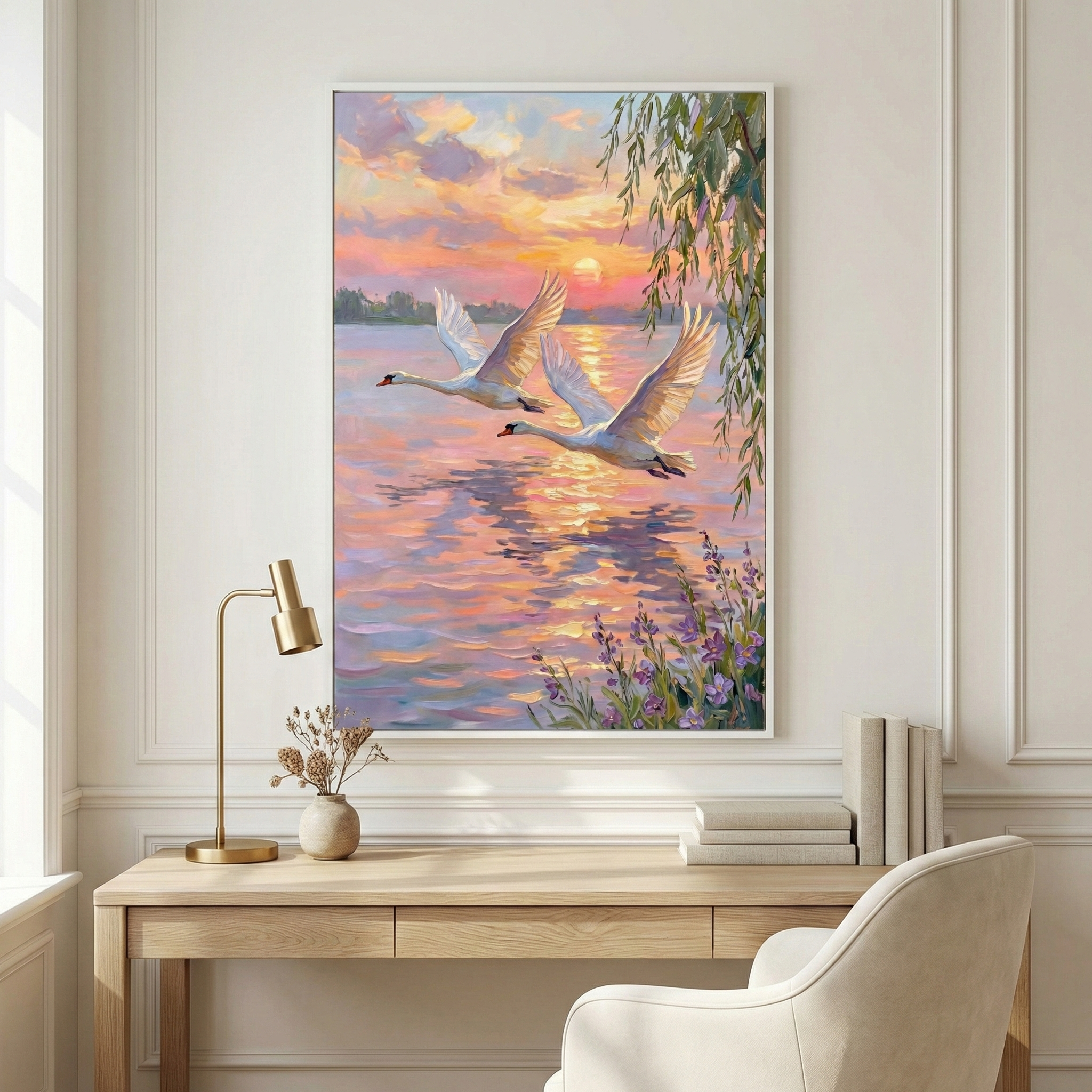 A vertical framed piece hangs on a white wall above a wooden desk. It depicts two white swans in flight over a shimmering lake during a sunset. The sky and water are filled with vibrant pink, orange, and purple hues. The sun glows on the horizon, casting a golden light. Wispy branches and purple flowers frame the scene. Below, a neat office space features a gold lamp, a vase with dried flowers, and books, creating a serene and elegant atmosphere.
