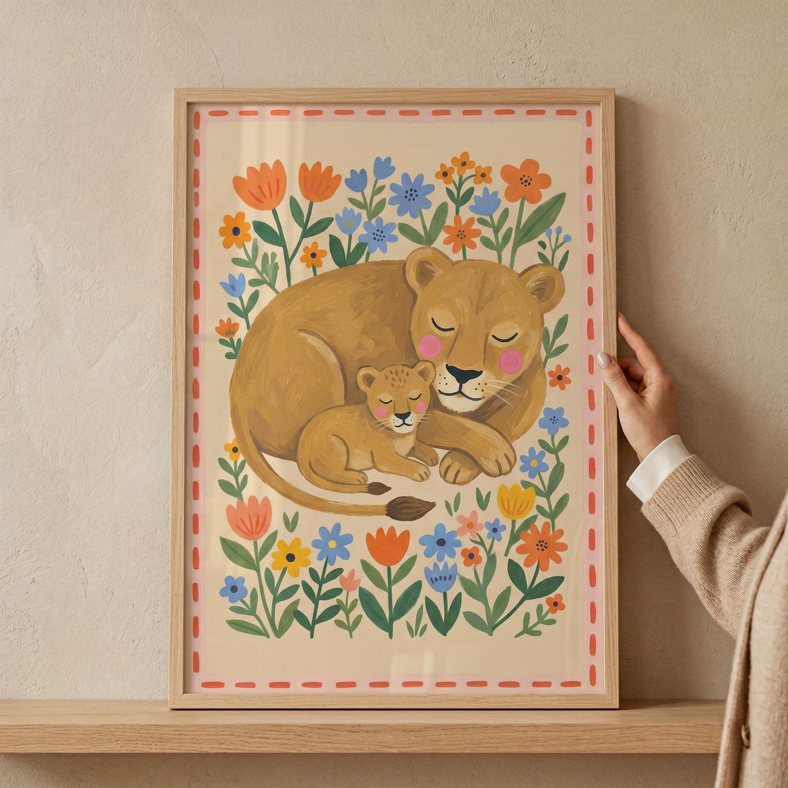 Framed folk art floral lioness artwork shows a mother lion and cub cuddling amidst vibrant flowers, held by a hand.