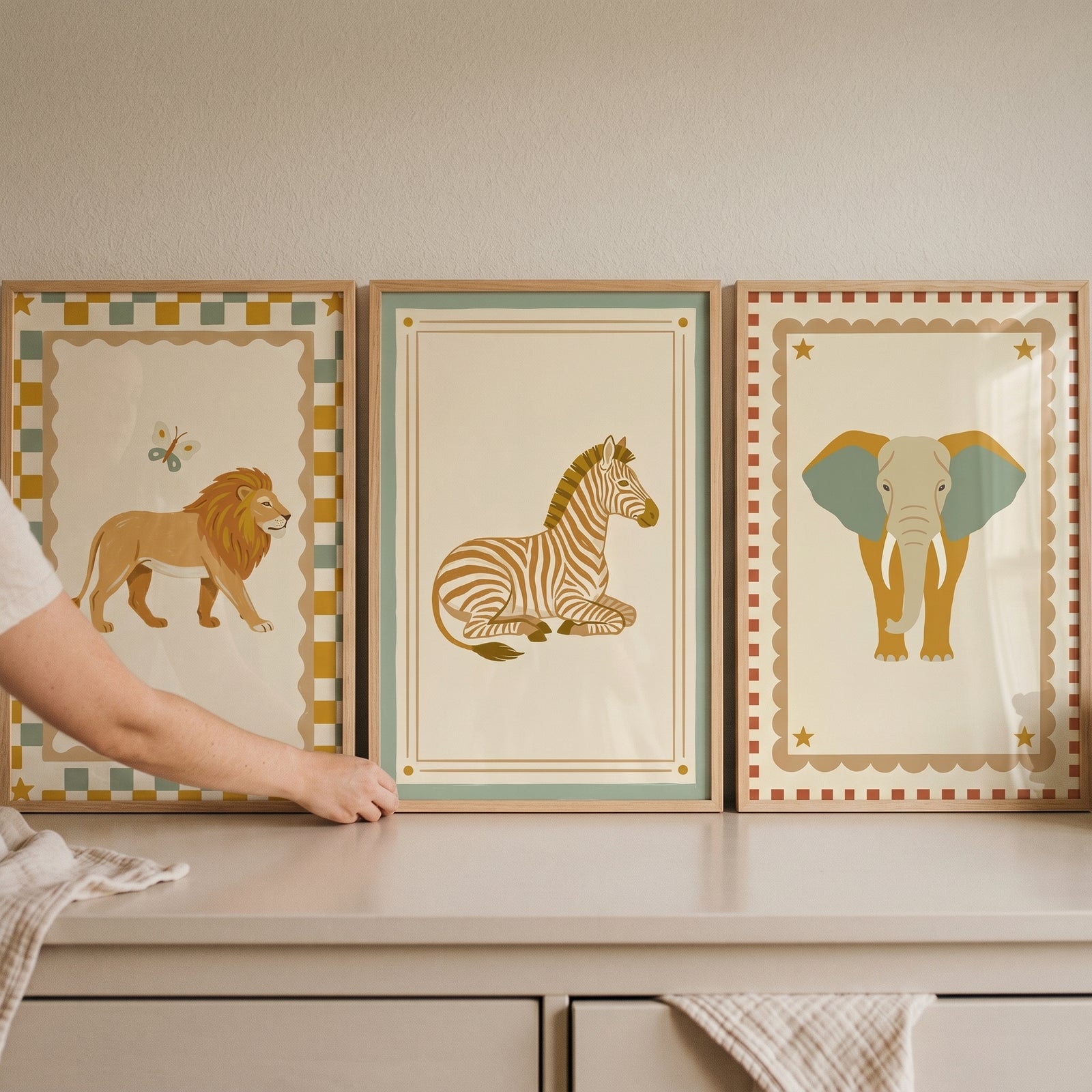 A hand adjusts a folk style safari animal wall art set of 3 featuring a lion, zebra, and elephant on a white dresser.