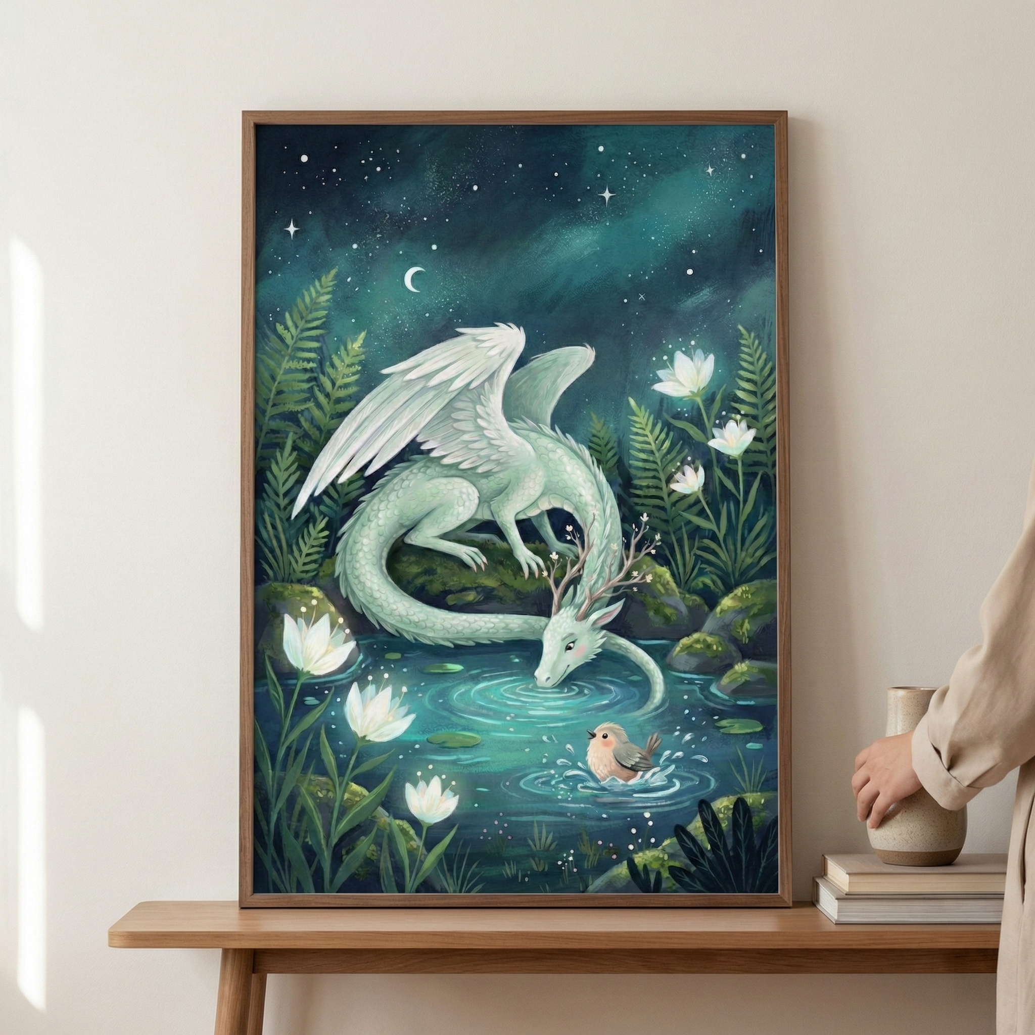 A whimsical illustration depicts a pale, mint-green dragon with large feathered wings and branching antlers drinking from a glowing pool of water. A small, round bird floats on the water's surface near the dragon's snout. The scene is set in a magical forest at night, surrounded by glowing white water lilies, lush ferns, and mossy rocks. Above, a dark teal sky is filled with stars and a crescent moon. The framed artwork is displayed on a wooden table.