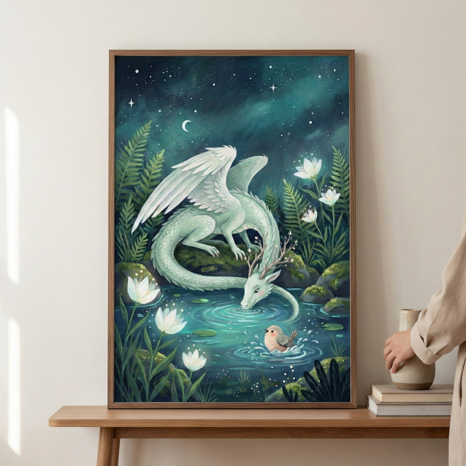 A whimsical illustration depicts a pale, mint-green dragon with large feathered wings and branching antlers drinking from a glowing pool of water. A small, round bird floats on the water's surface near the dragon's snout. The scene is set in a magical forest at night, surrounded by glowing white water lilies, lush ferns, and mossy rocks. Above, a dark teal sky is filled with stars and a crescent moon. The framed artwork is displayed on a wooden table.