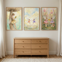 Three framed watercolor illustrations of fairies hang on a plain white wall above a light-colored wooden dresser. The artwork on the left features a fairy leaning against a large mushroom while reading. The central illustration shows two fairies dancing in a circle above a ring of small mushrooms. The piece on the right depicts two fairies sitting on mushrooms in a grassy field. The six-drawer dresser has simple round knobs. White curtains frame the scene, and a textured rug lies on the floor.