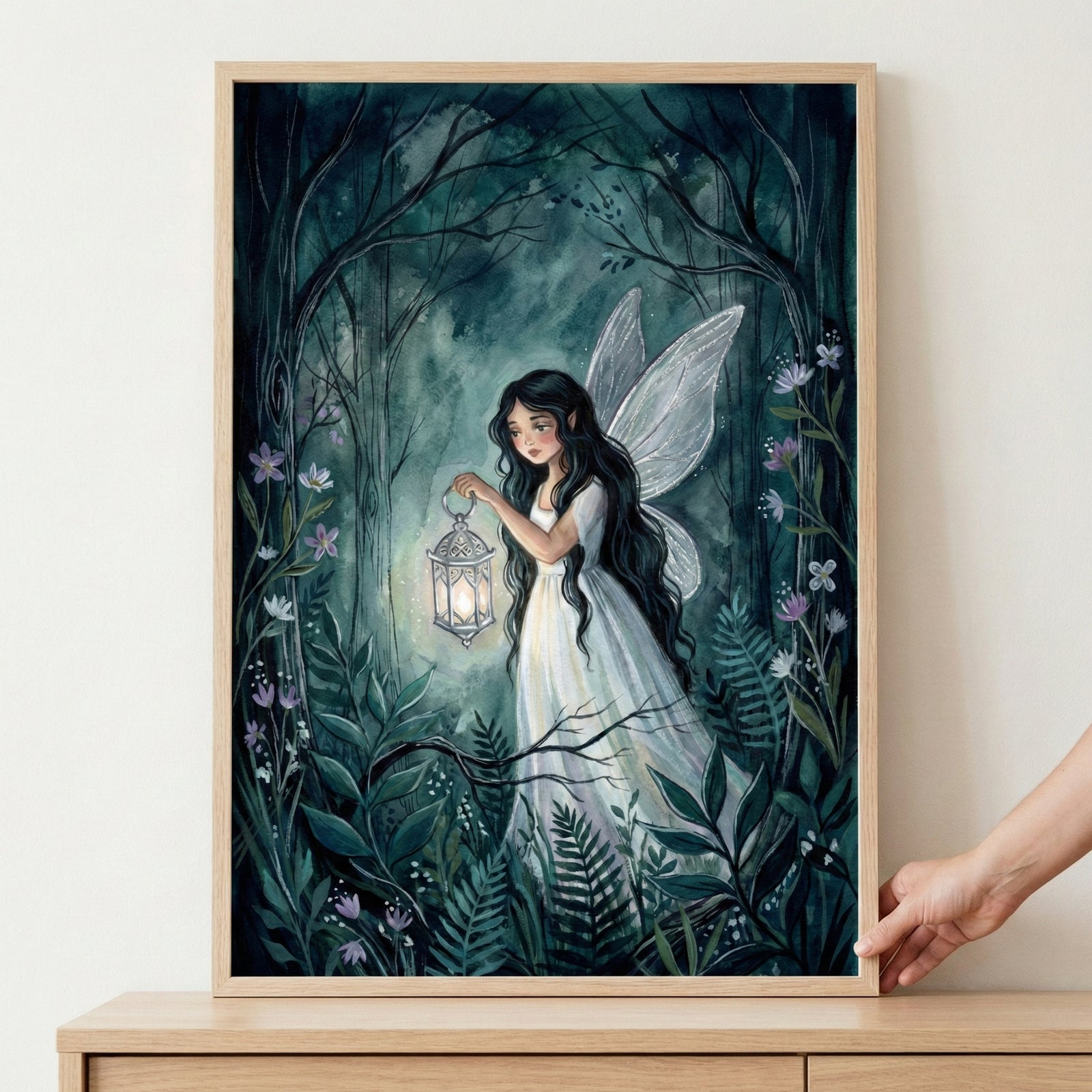 A framed illustration of a fairy with long dark hair and translucent wings walking through a dark, mystical forest. She wears a flowing white dress and holds a glowing lantern that illuminates the surrounding ferns and small purple flowers. The forest is rendered in deep shades of teal and green, with gnarled trees arching overhead. A hand is visible on the right side, holding the edge of the light-colored wooden frame.