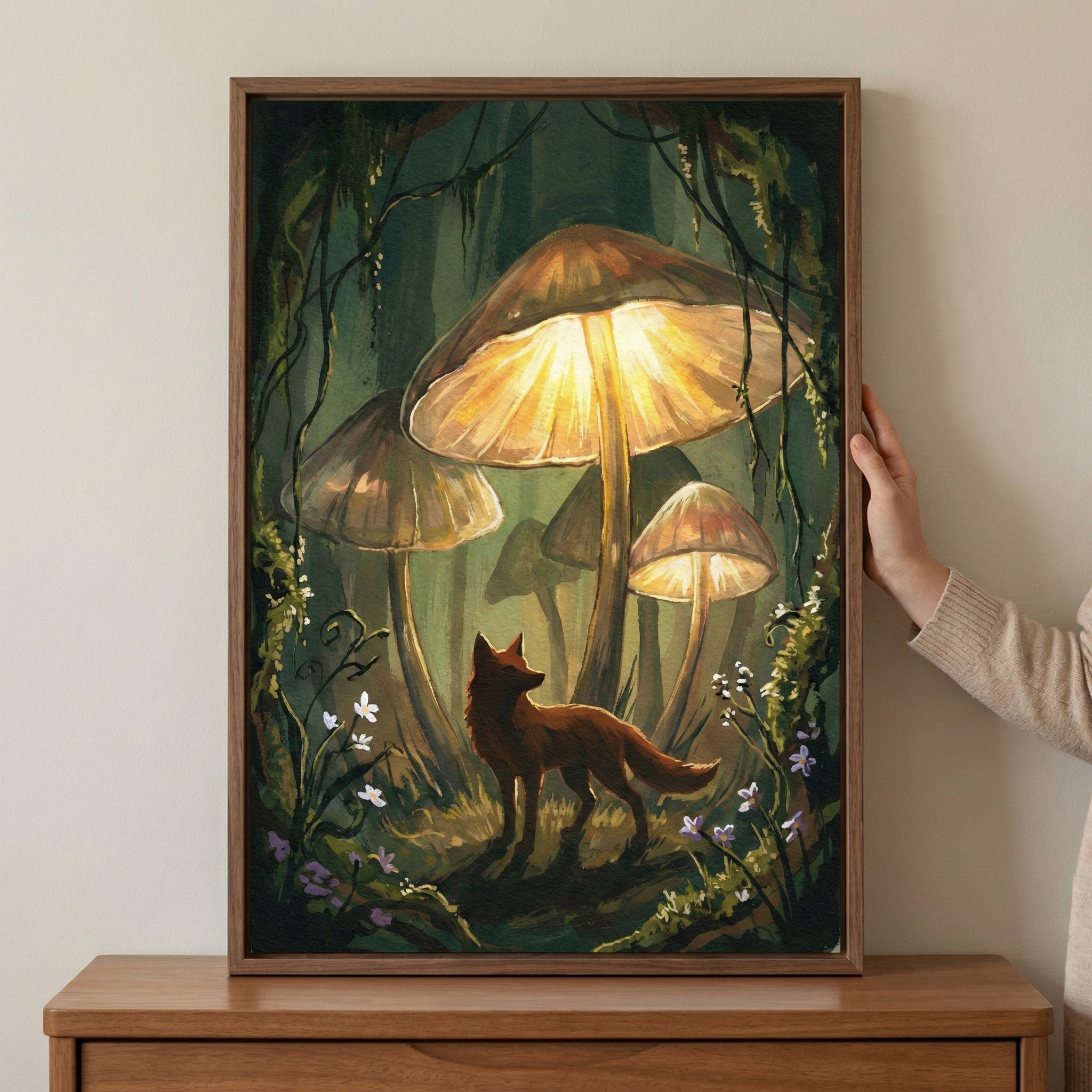 A framed vertical illustration of a red fox standing beneath oversized, glowing mushrooms in a dark forest. The central mushroom radiates a warm golden light, illuminating the fox and the surrounding foliage of vines and small wildflowers. A hand holds the side of the wooden frame, which rests on a wooden surface against a neutral wall.