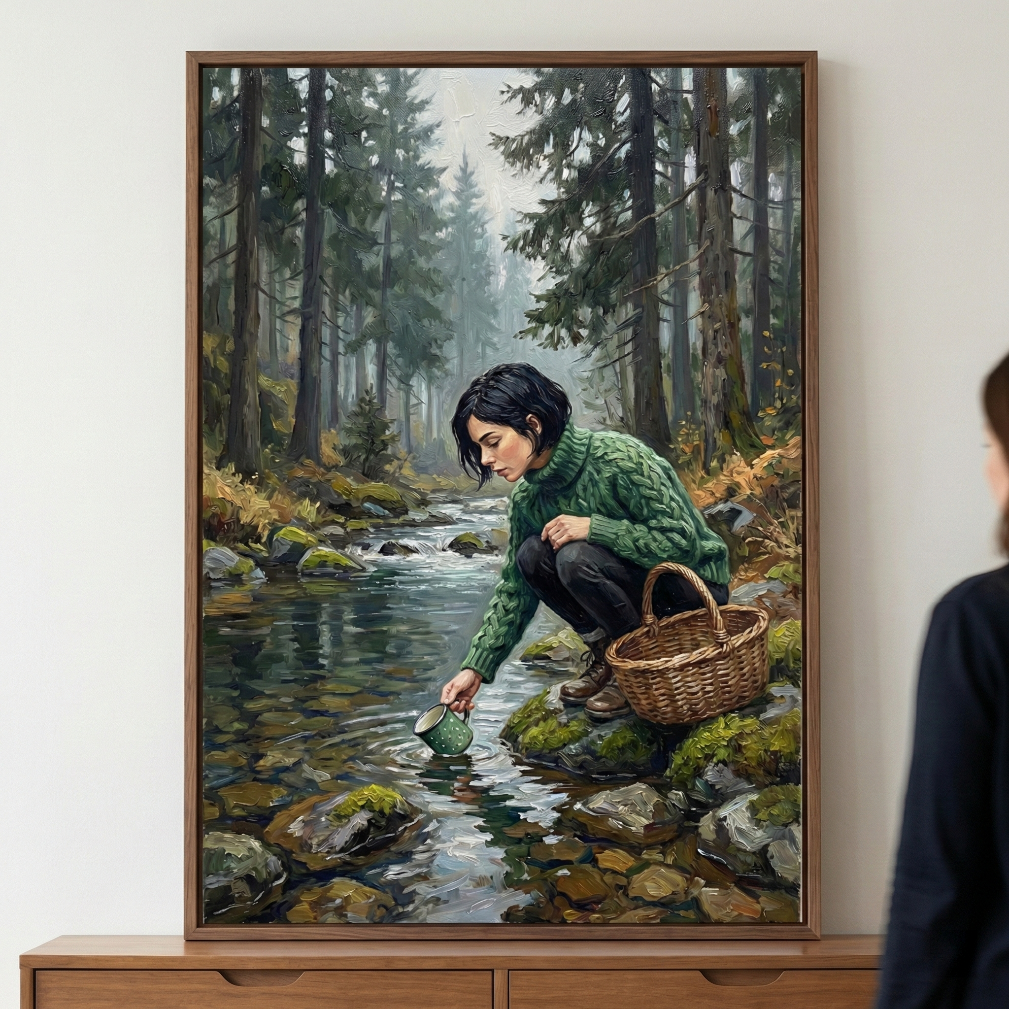 A vertical framed artwork depicts a woman with short dark hair crouching by a clear, rocky stream in a dense pine forest. She wears a chunky green cable-knit sweater and dark trousers, reaching down to dip a small green mug into the flowing water. Beside her on a moss-covered rock sits a woven wicker basket. The scene is rendered with expressive brushstrokes, highlighting the textures of the knitwear, the rippling water, and the forest floor.