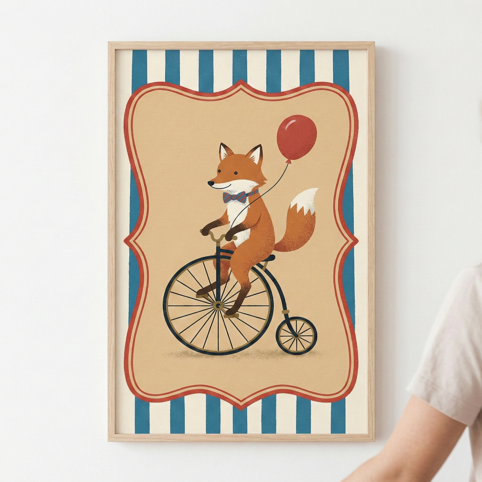 A light wood-framed vertical illustration of a whimsical cartoon fox riding a vintage-style high-wheel bicycle. The fox wears a small striped bowtie and holds a single red balloon. This central scene is enclosed within a decorative red and cream scalloped border. The overall background consists of vertical blue and cream stripes. The frame is mounted on a plain white wall, with a person's shoulder and arm partially visible in the lower right corner.